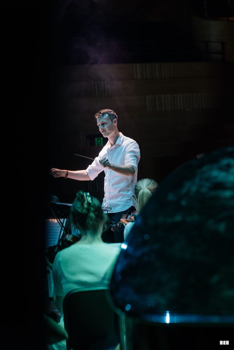 A conductor leads an orchestra during a performance, illuminated by stage lighting with audience members visible in the foreground.