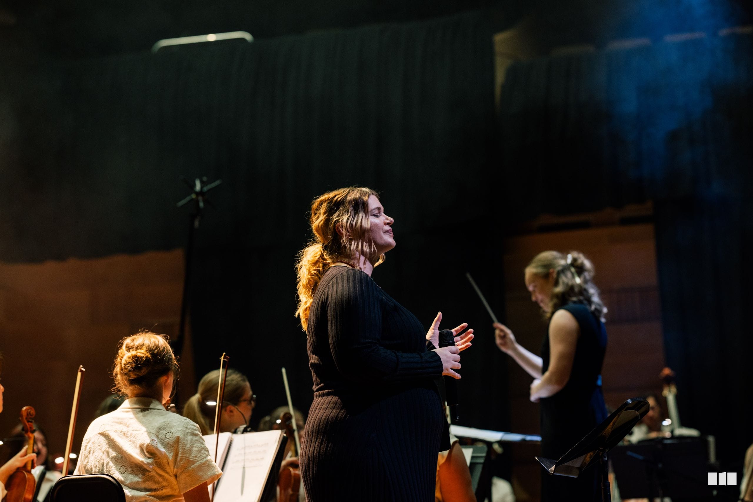 A conductor leads an orchestra during a performance, with string players visible in the foreground and ensemble members focused on the music.