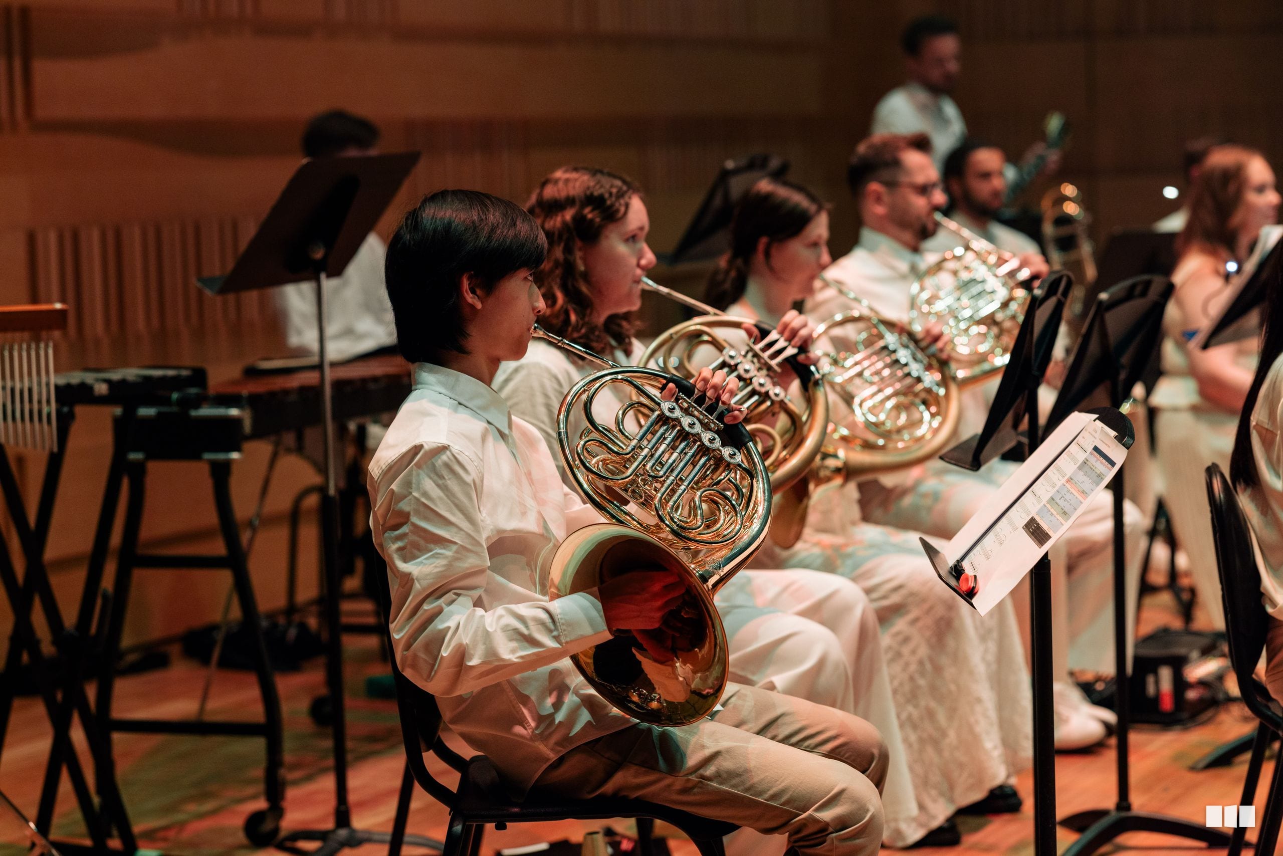 French horn players seated in the orchestra section during a rehearsal or performance, reading from sheet music.