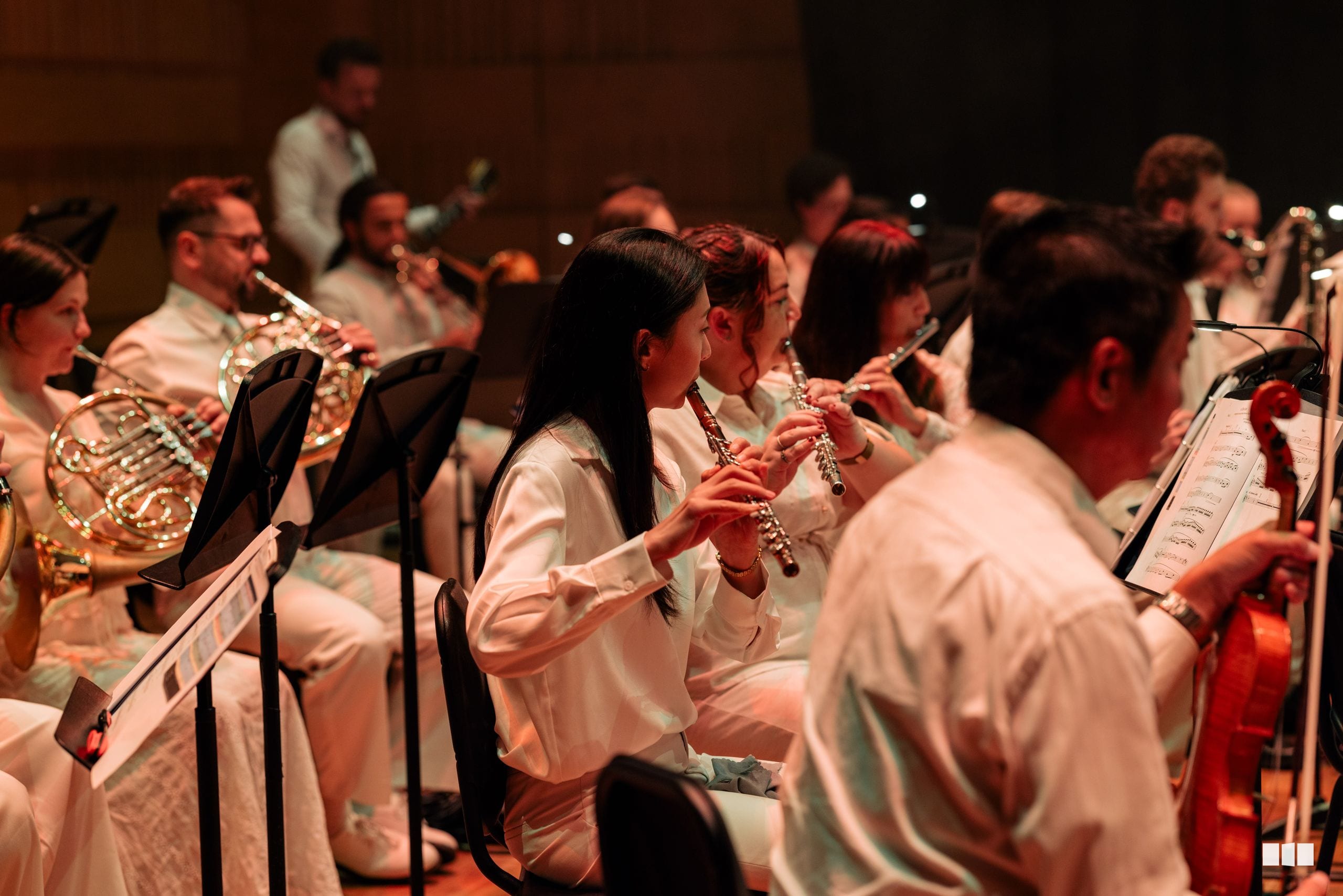 Wind and brass musicians perform together during an orchestral concert, with flute players prominently featured in the foreground.