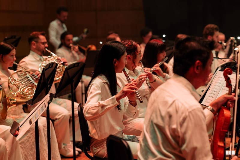 Wind and brass musicians perform together during an orchestral concert, with flute players prominently featured in the foreground.