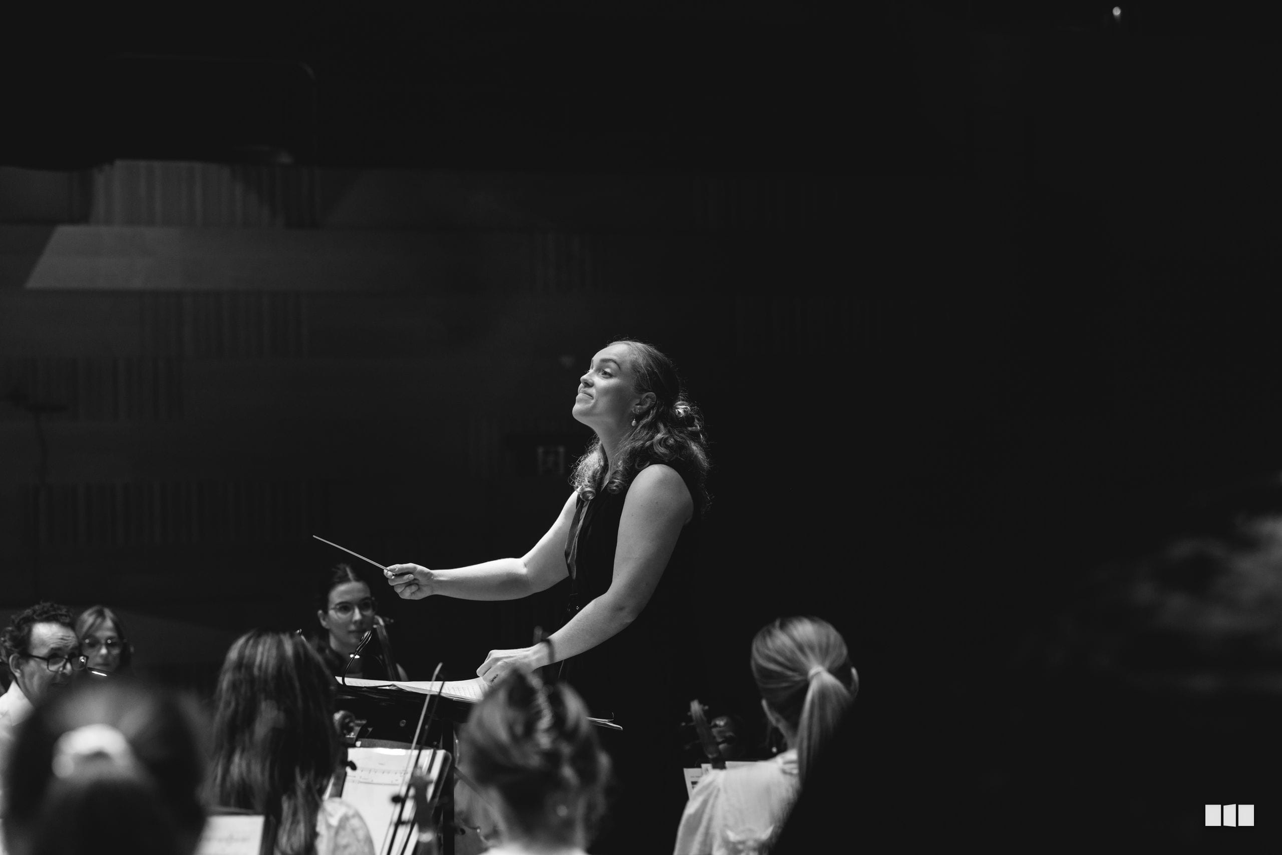 A conductor leads an orchestra with an expressive upward gesture, captured in black and white during a performance.