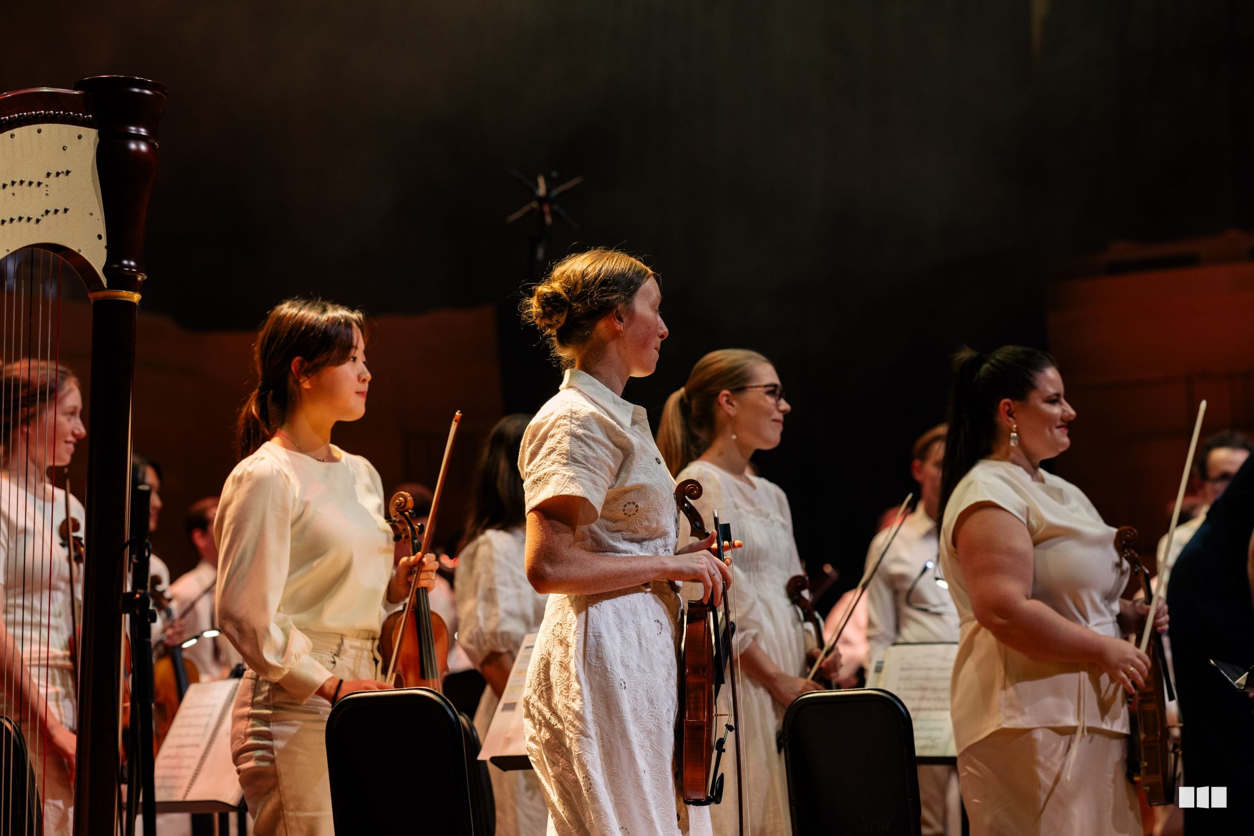 String players in white attire stand in profile during an orchestral performance on stage.