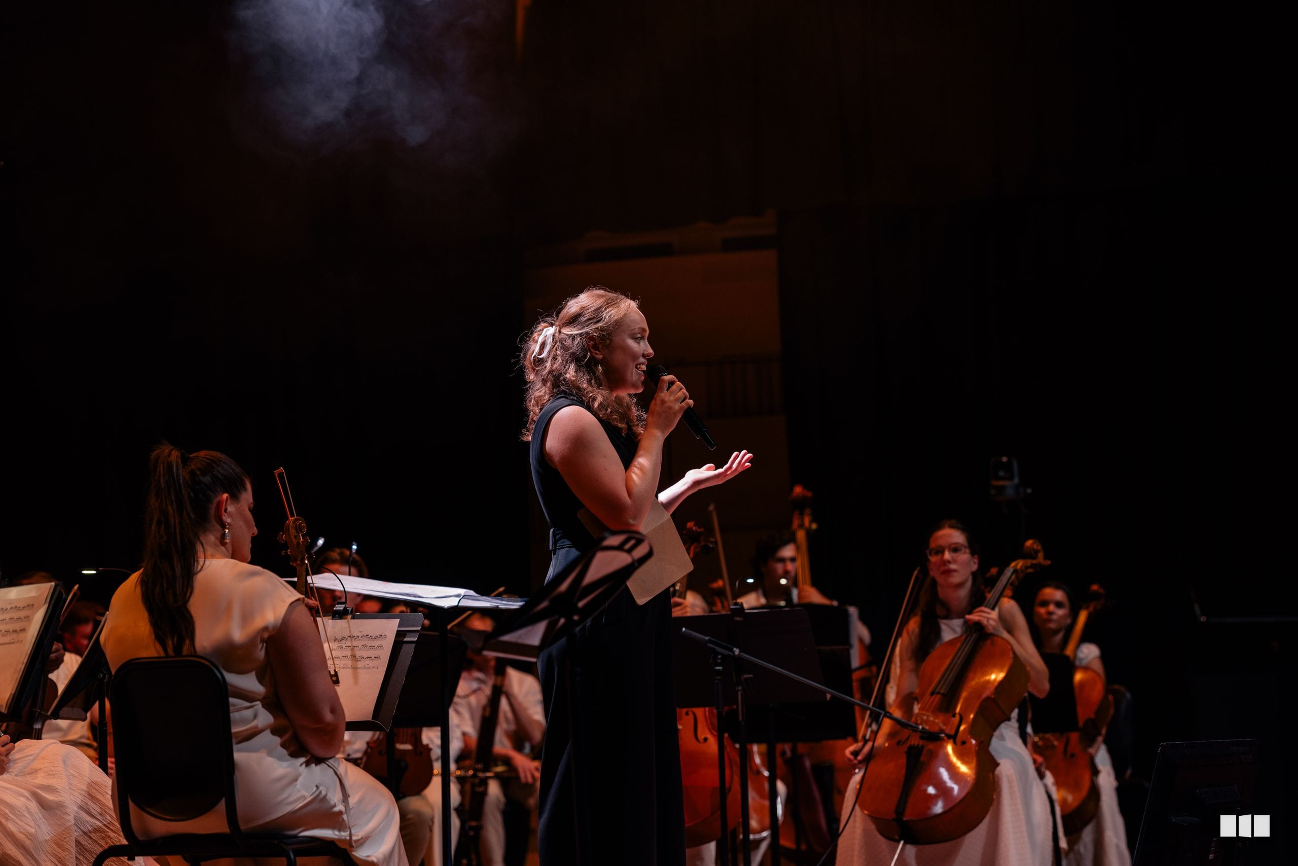 A soprano soloist performs with microphone while accompanied by string musicians on stage in warm stage lighting.