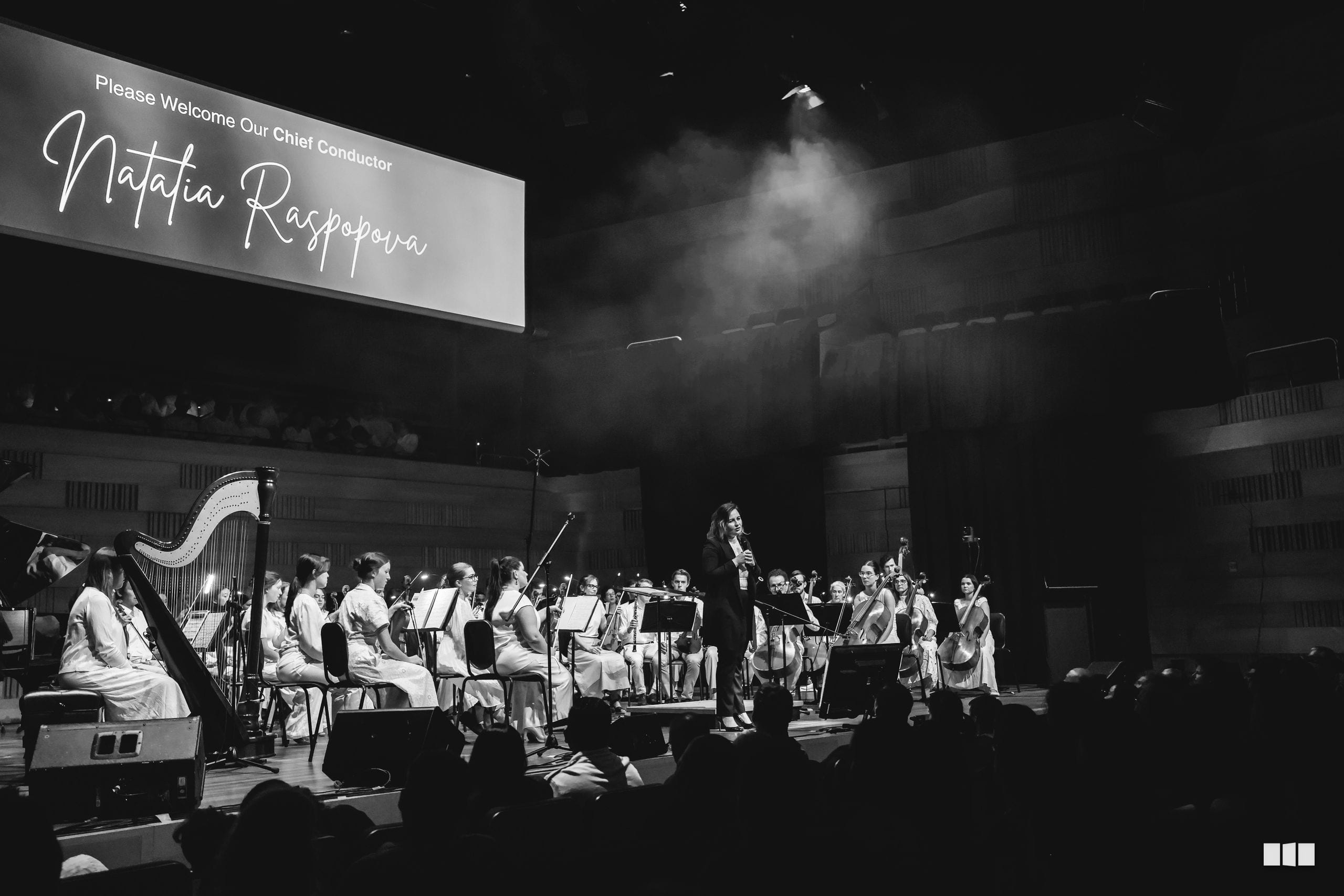 A symphony orchestra performs on stage during the welcome of Chief Conductor Natalia Raspopoura, with musicians in formal white attire arranged across the stage.
