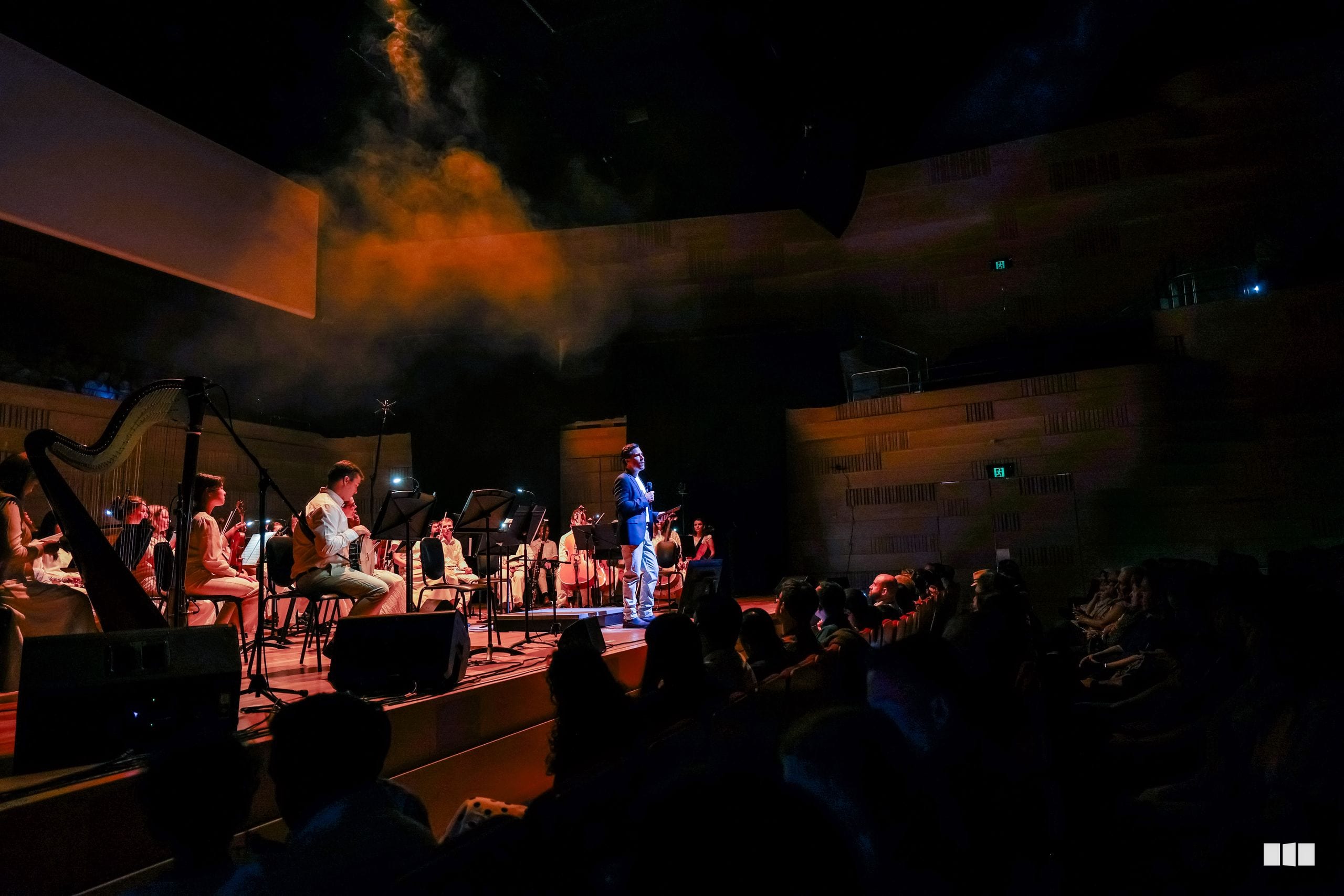A conductor leads an orchestra and ensemble in an outdoor evening performance in front of an apartment building, with audience members seated in the foreground.