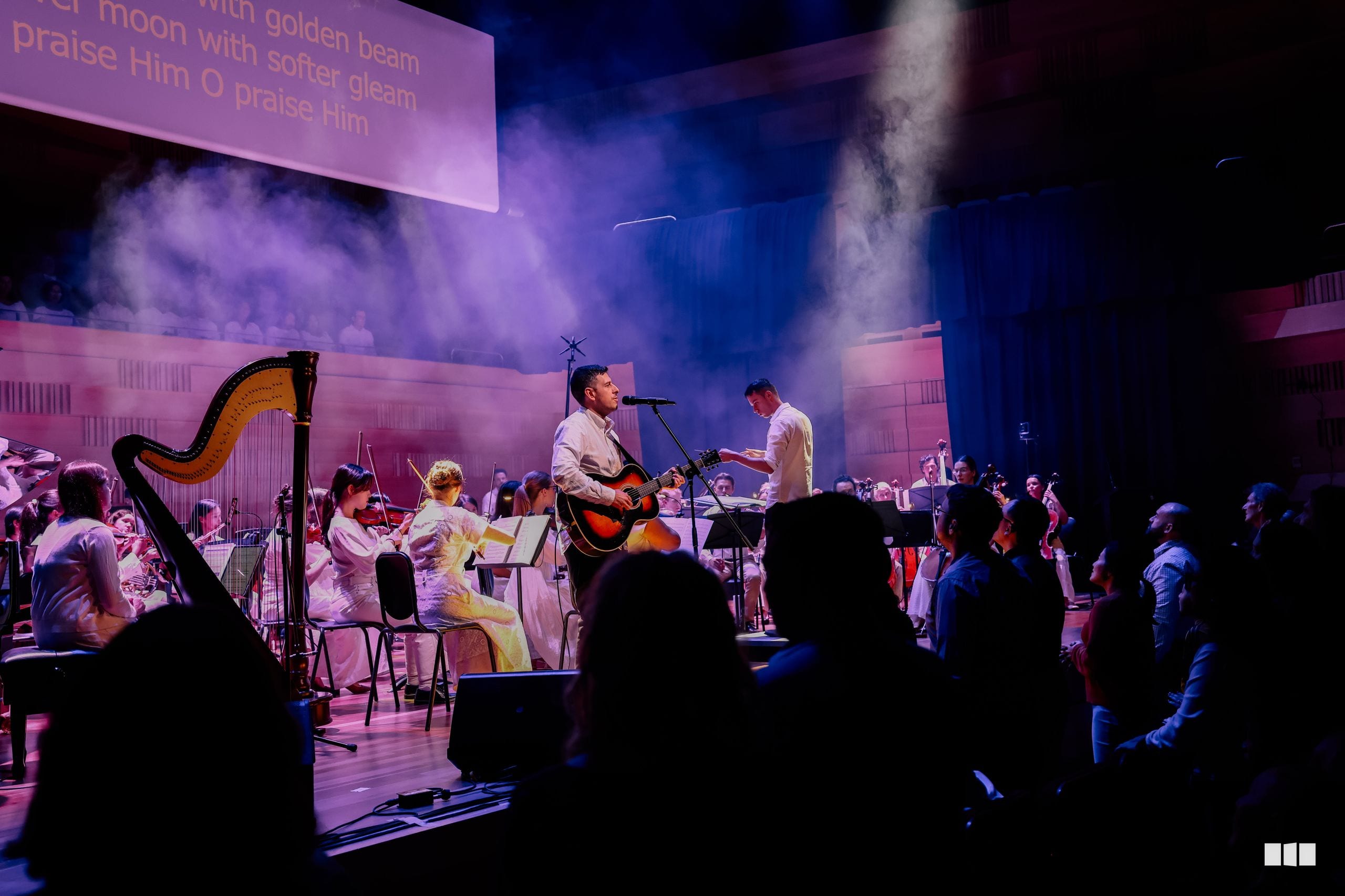 A male soloist performs with acoustic guitar alongside orchestra and choir members on stage, bathed in purple and blue lighting with atmospheric effects.