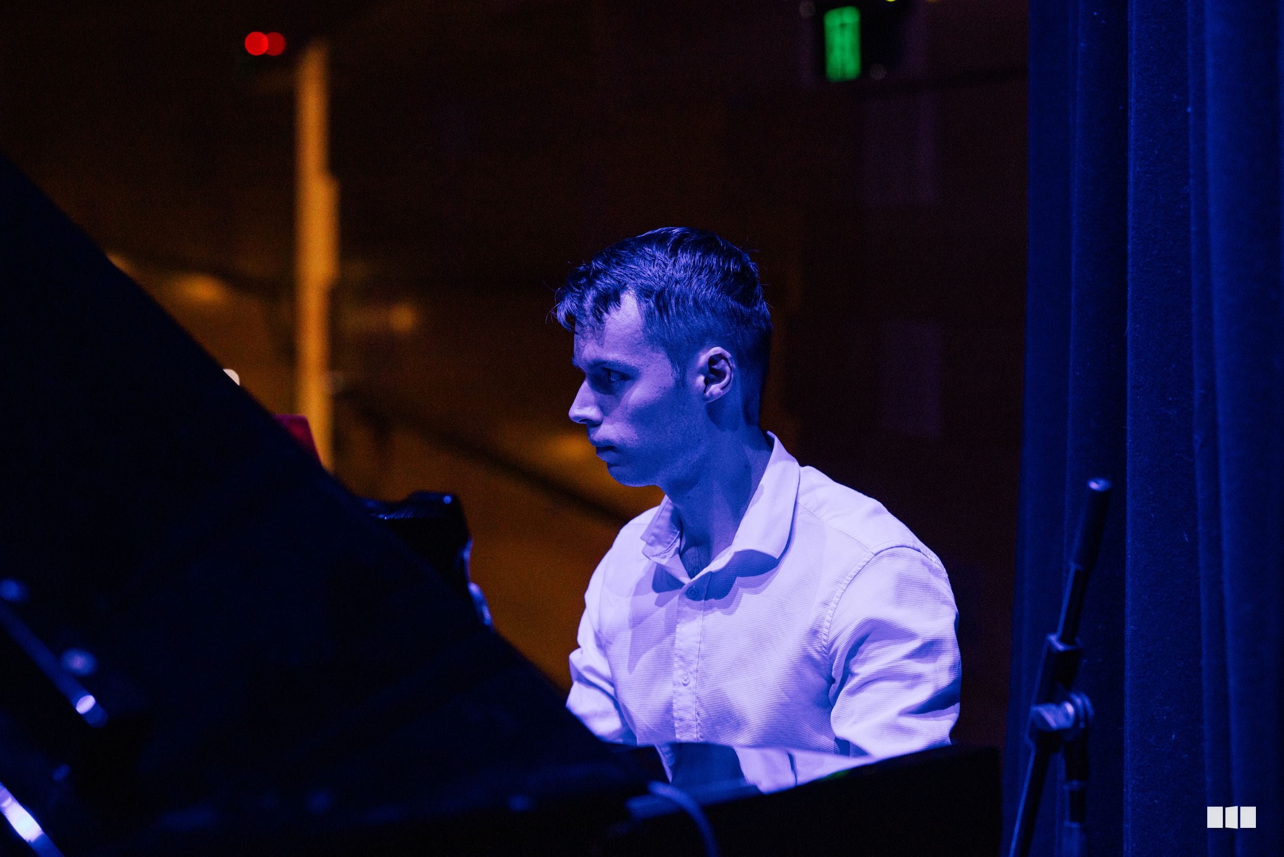 A pianist in a white shirt performs at the keyboard during an evening concert, illuminated by blue stage lighting.