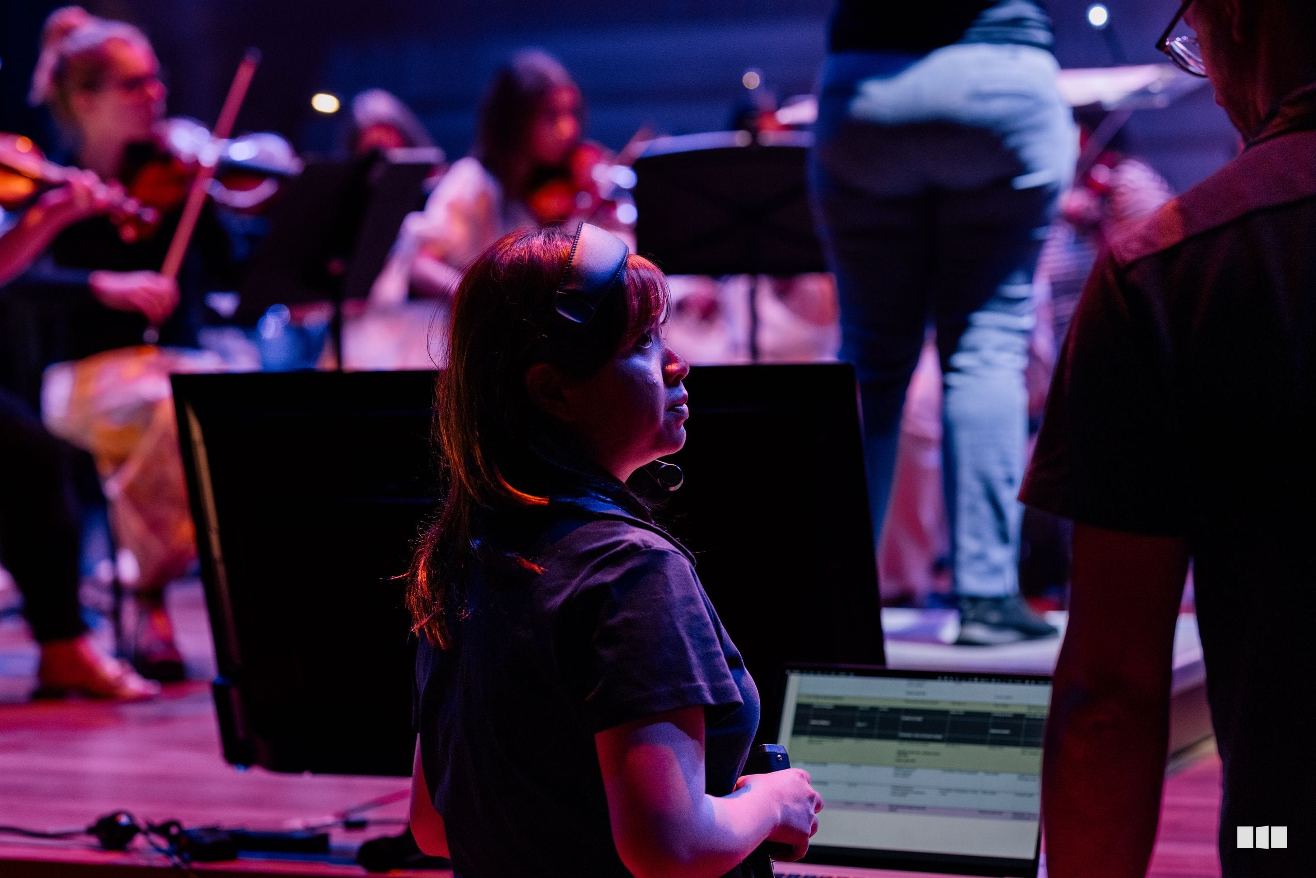 A young performer stands engaged during an orchestral rehearsal or performance with musicians and instrumental equipment visible in the atmospheric stage lighting.