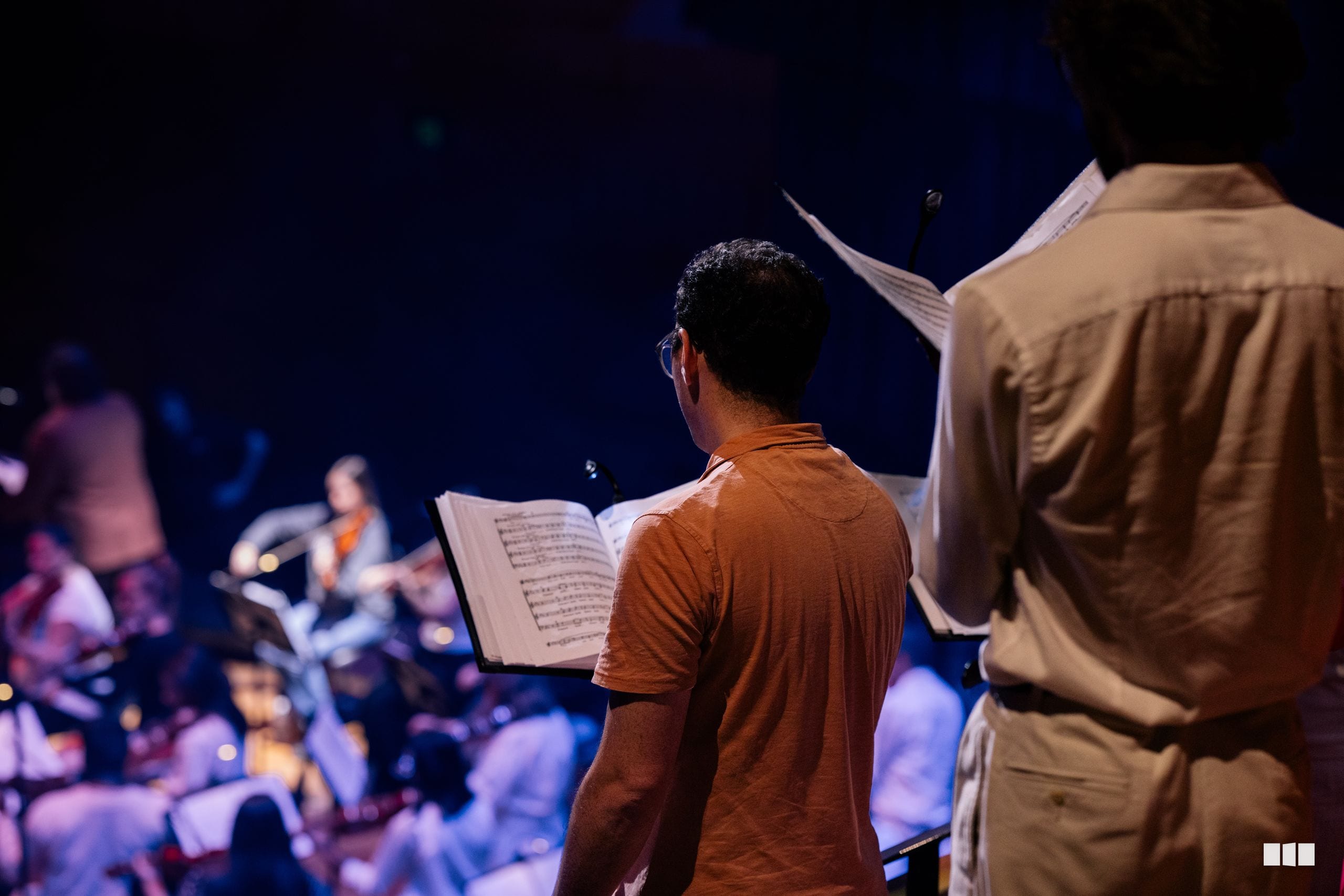 A conductor in an orange shirt holds sheet music while leading the orchestra during a performance in a concert hall.