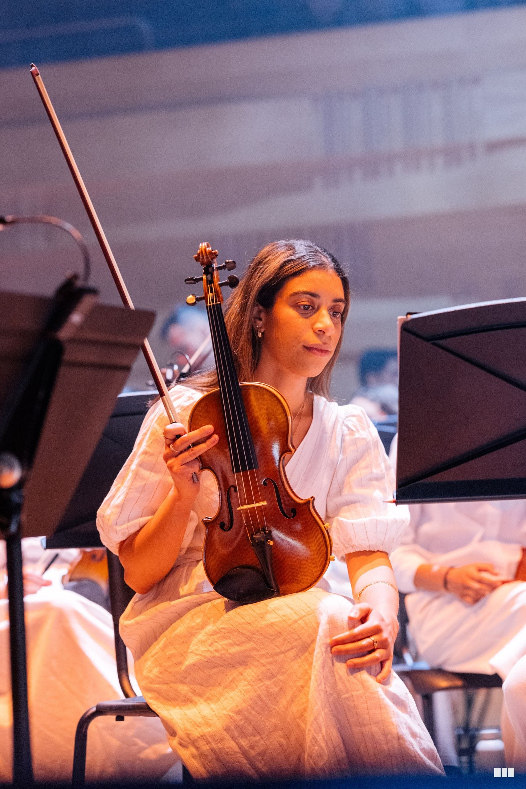 A violinist in white attire performs with bow raised during an orchestral rehearsal or concert.