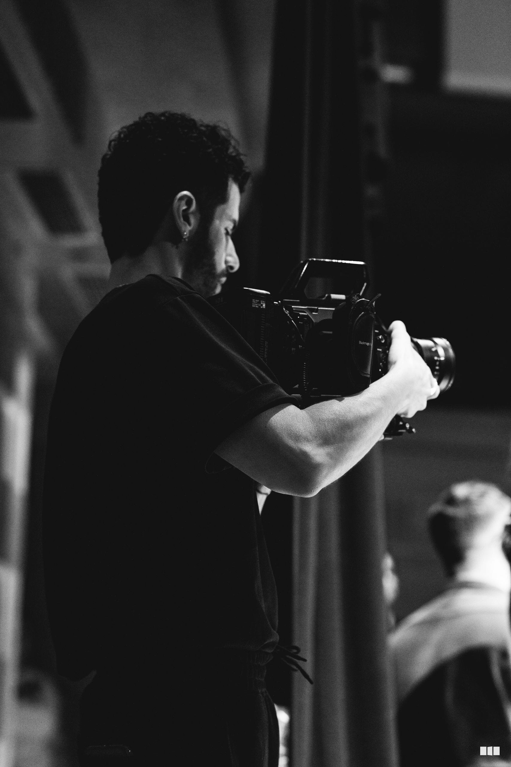 A bearded cameraman in black attire holds a professional video camera while filming indoors.