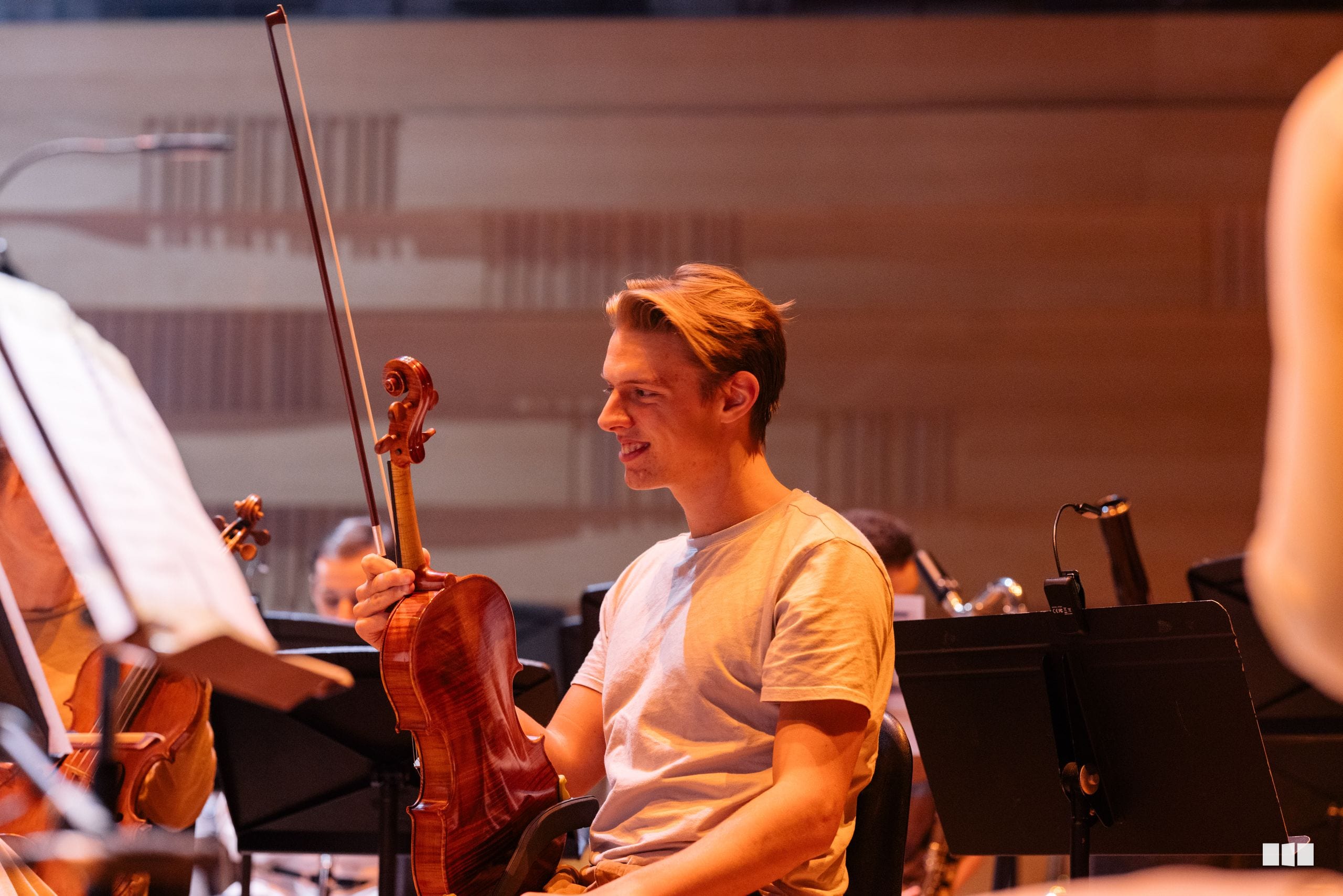 A young violinist smiles while holding his instrument during an orchestral rehearsal or performance.