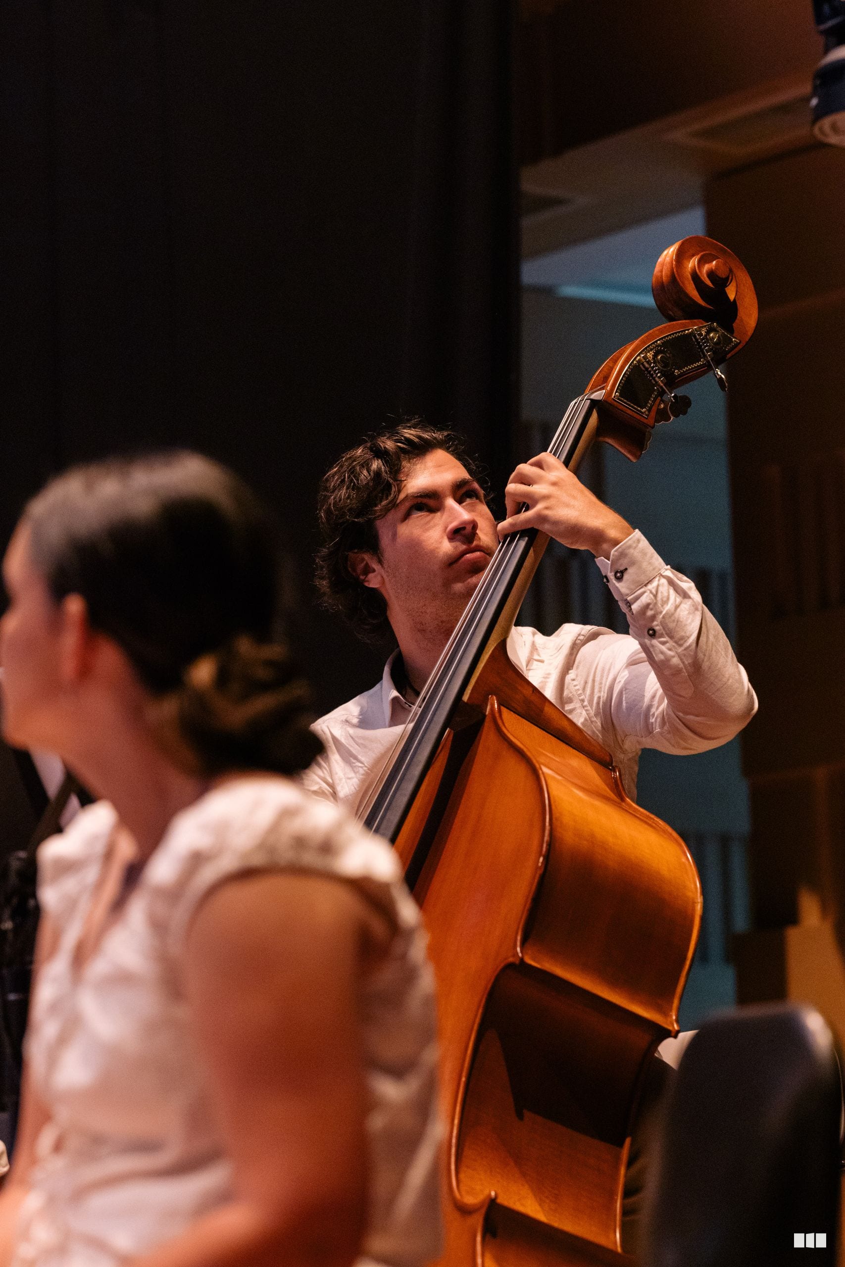 A cellist in white shirt performs with focused expression during an orchestral rehearsal or concert.