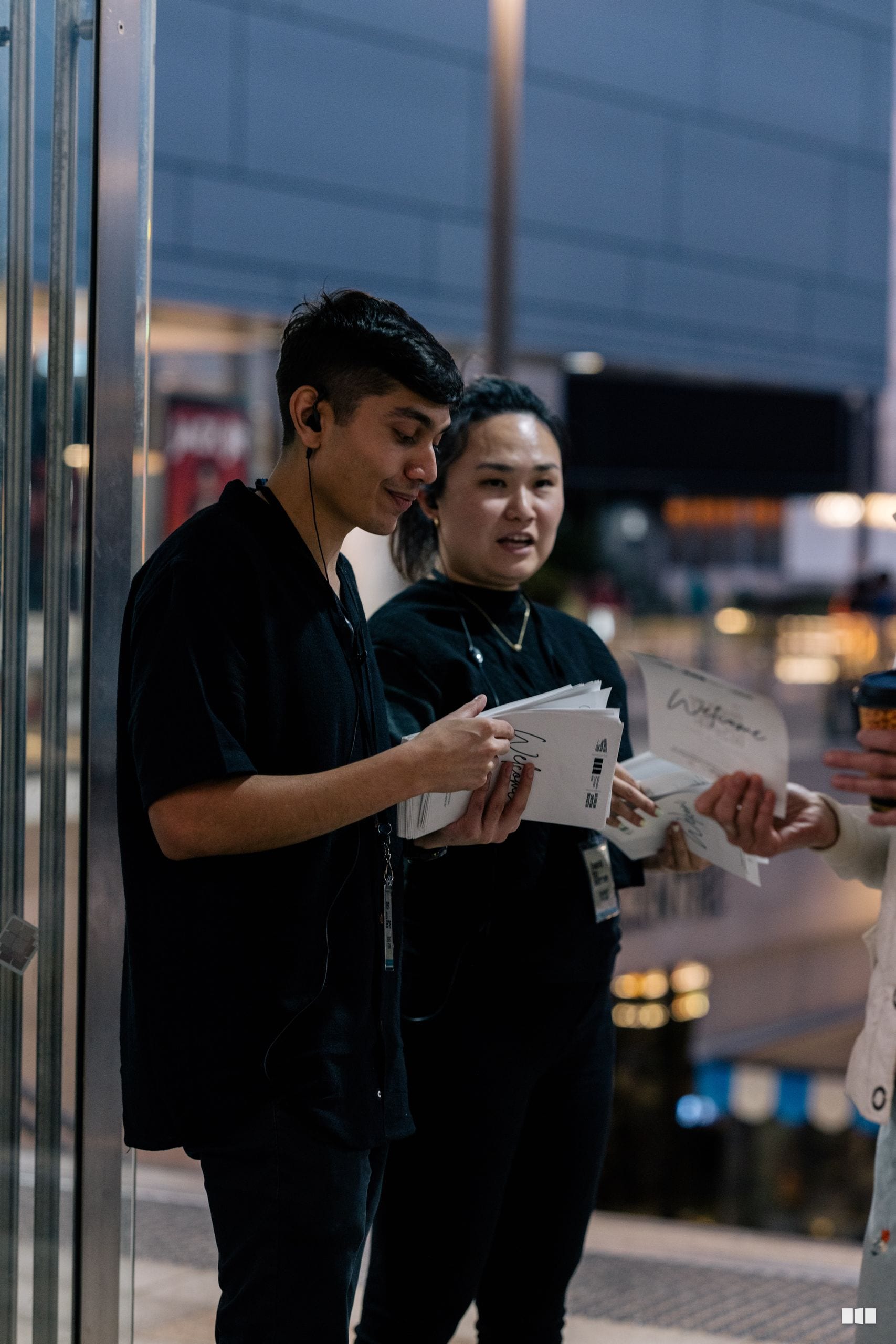 Two young people in black attire review documents together outside a venue during evening hours.