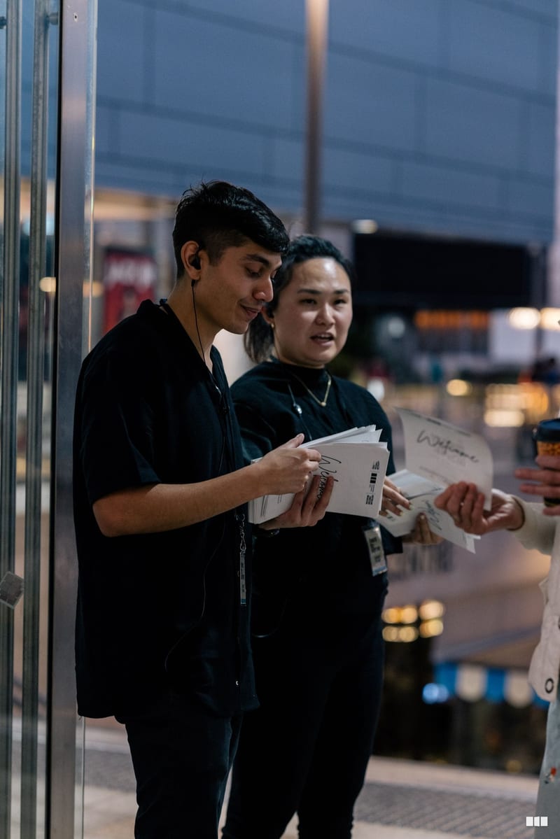 Two young people in black attire review documents together outside a venue during evening hours.