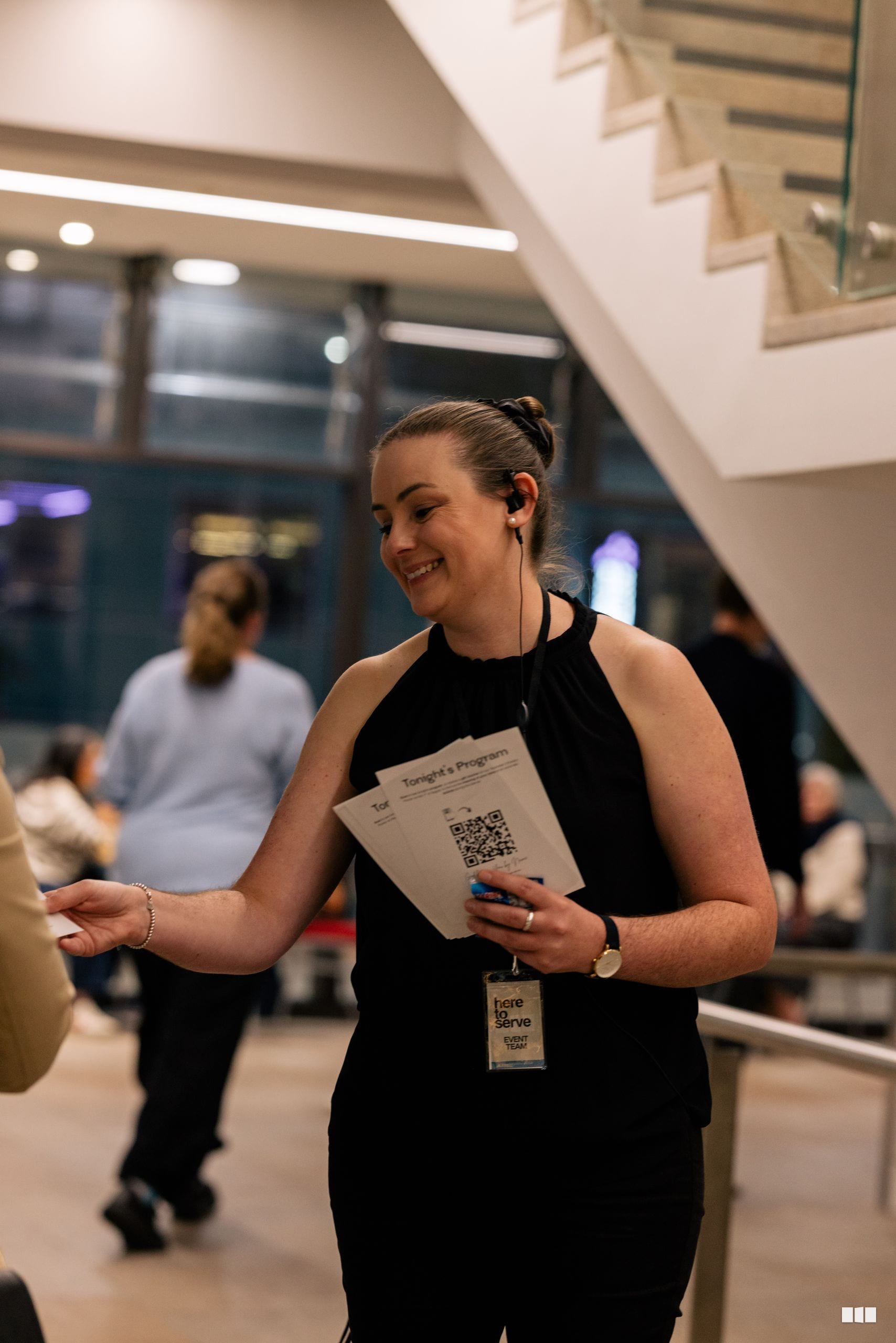 A smiling volunteer or staff member holding a program sheet with QR code at an indoor event venue.