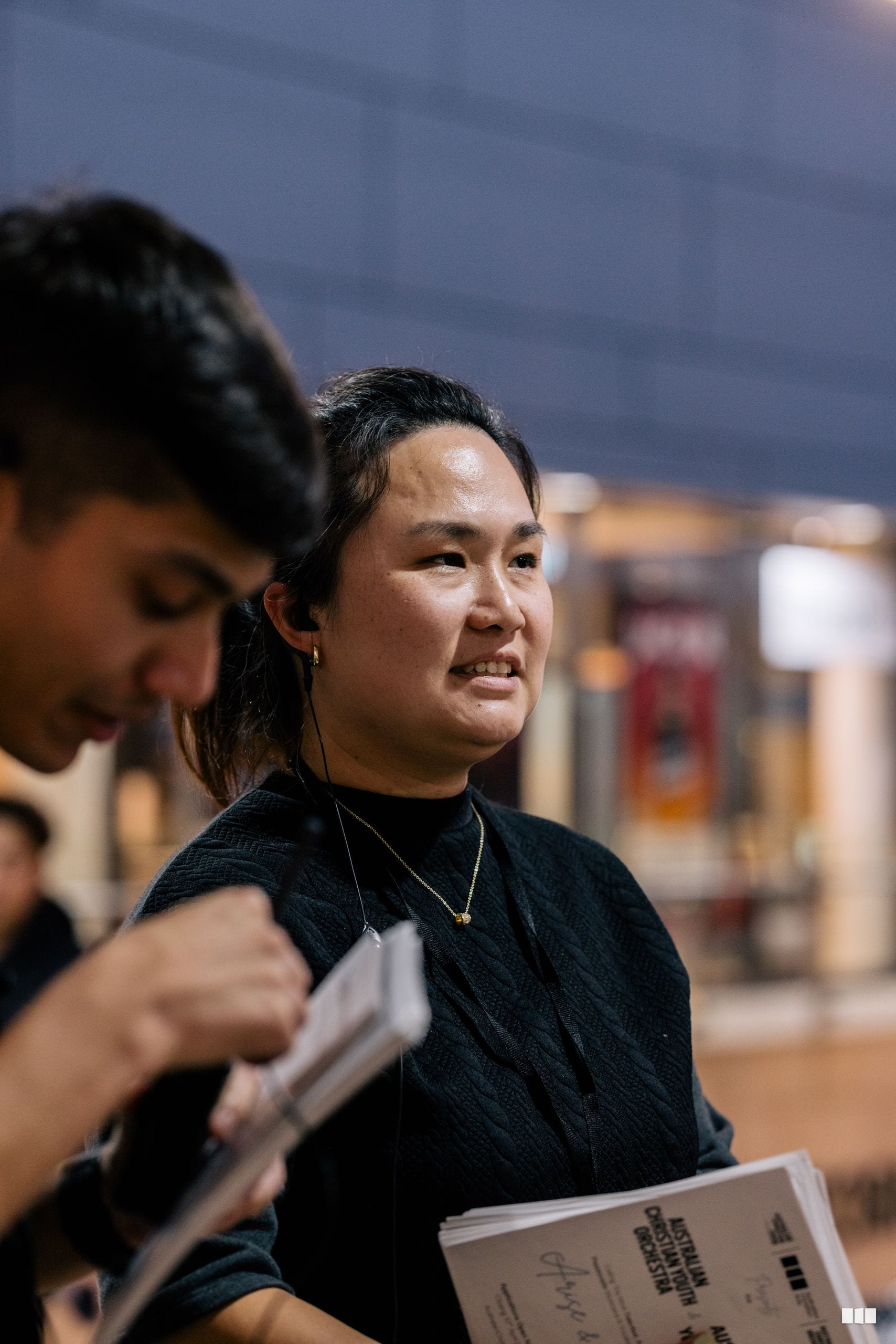A young woman holds printed materials while appearing to participate in or attend an event, with blurred activity visible in the background.