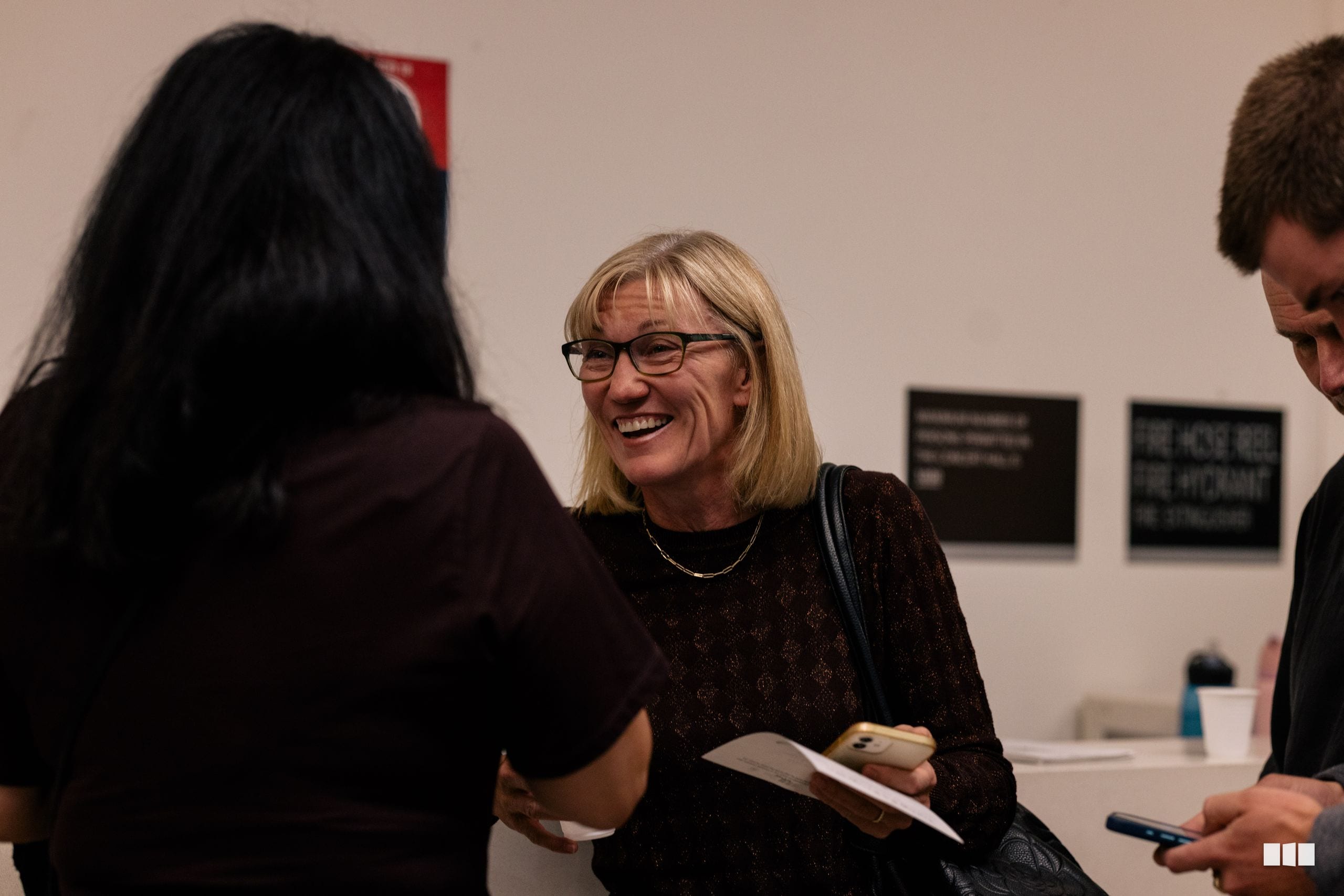 A woman in glasses smiles while conversing with attendees at an indoor event, holding a program and mobile phone.