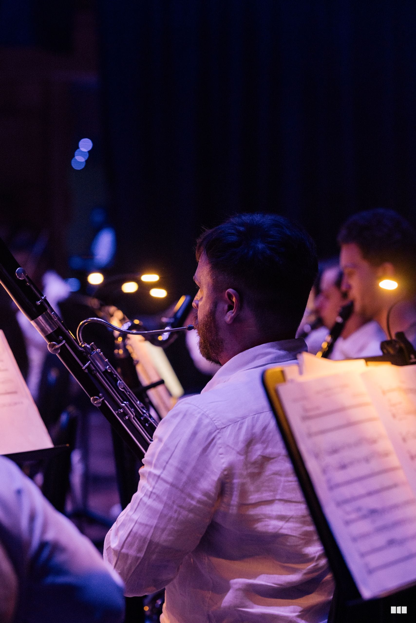 A woodwind musician plays saxophone during an evening orchestral performance, with sheet music visible and stage lighting in the background.