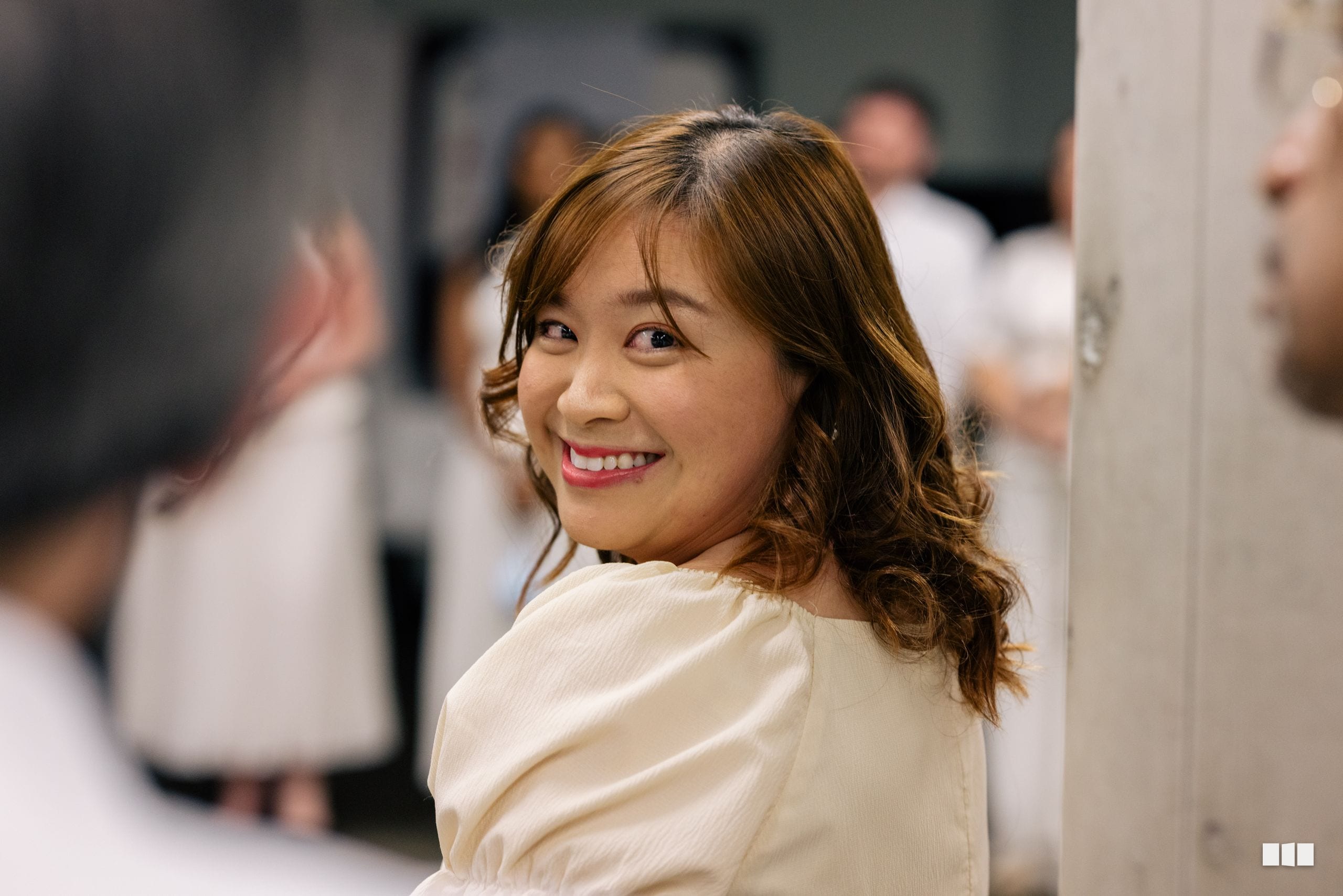 A woman in a cream-colored blouse smiles warmly at the camera during what appears to be a social gathering or event.