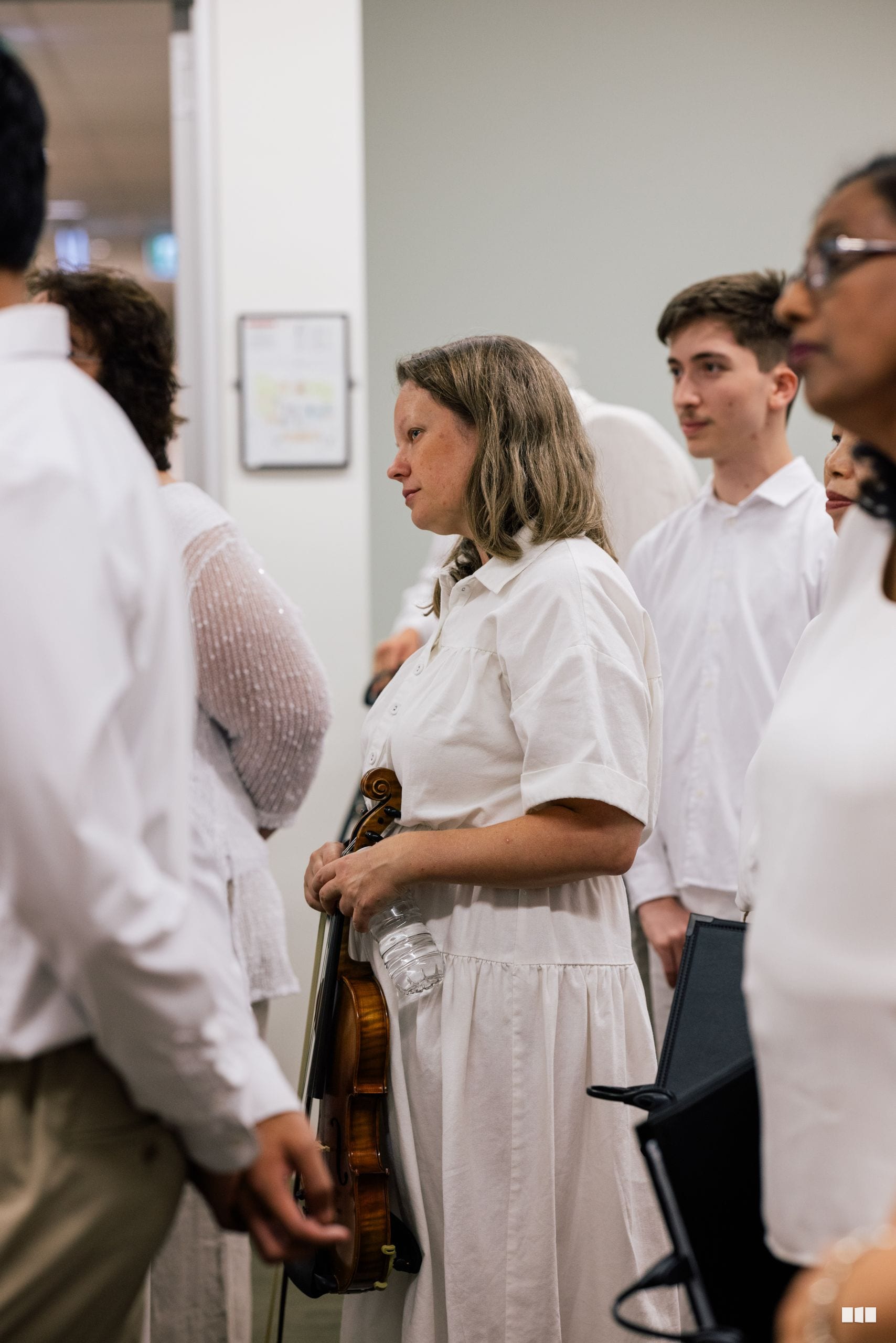 A violinist in white attire stands among other performers during a rehearsal or performance preparation in an indoor venue.