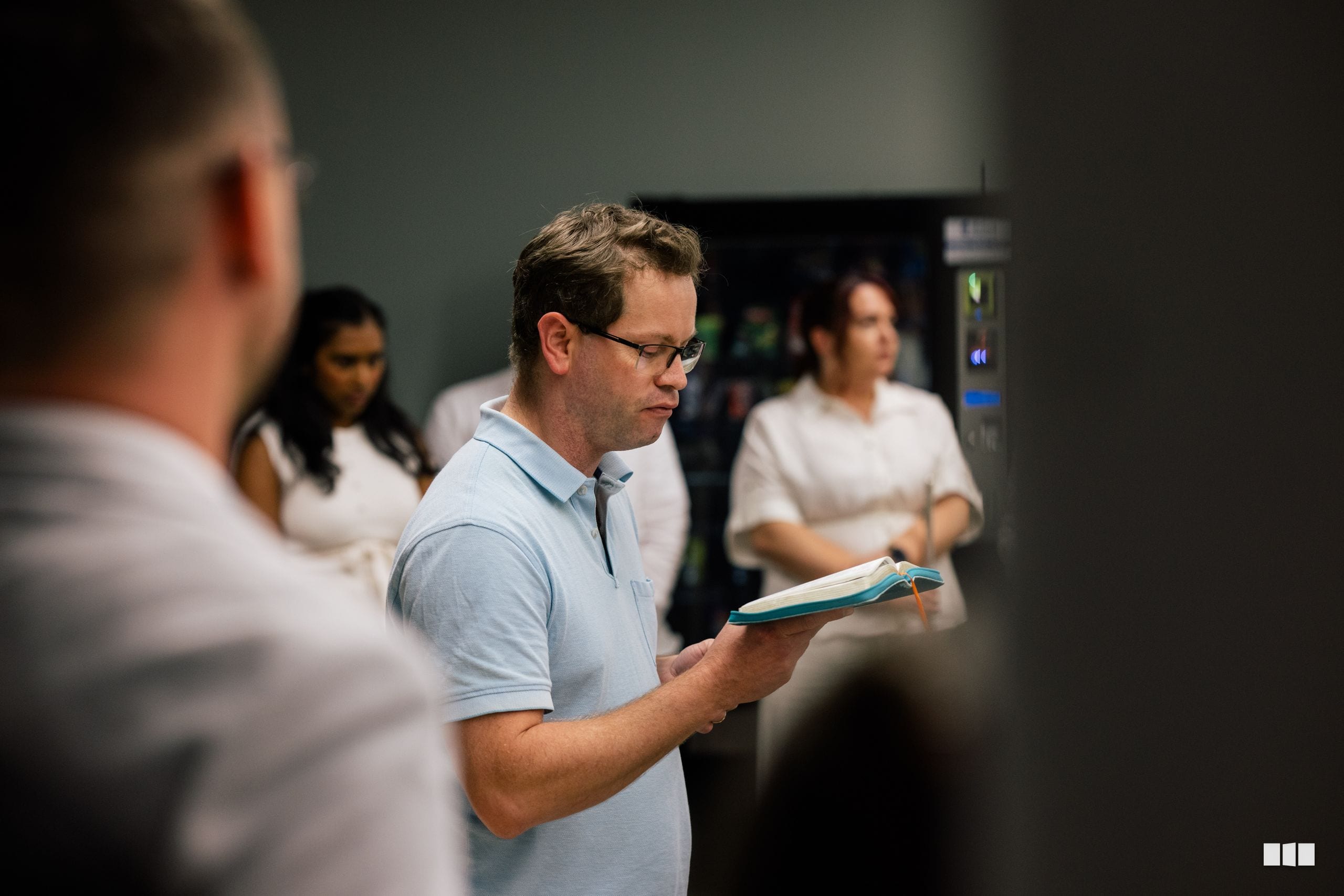 A man in a light blue polo shirt holds an open notebook while leading a group discussion in an indoor venue.