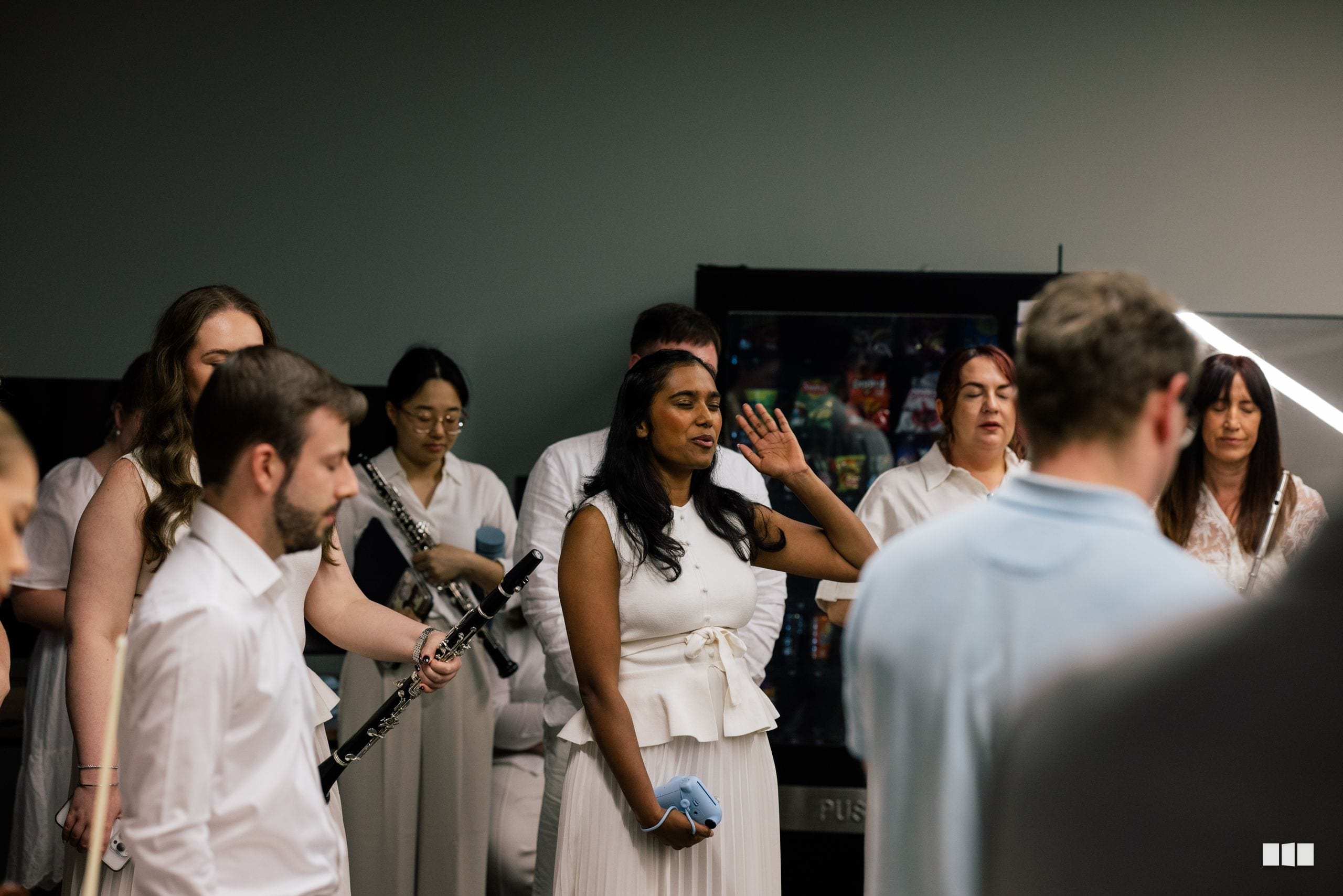 A choir member in white attire gestures expressively while rehearsing with ensemble members in a studio setting.