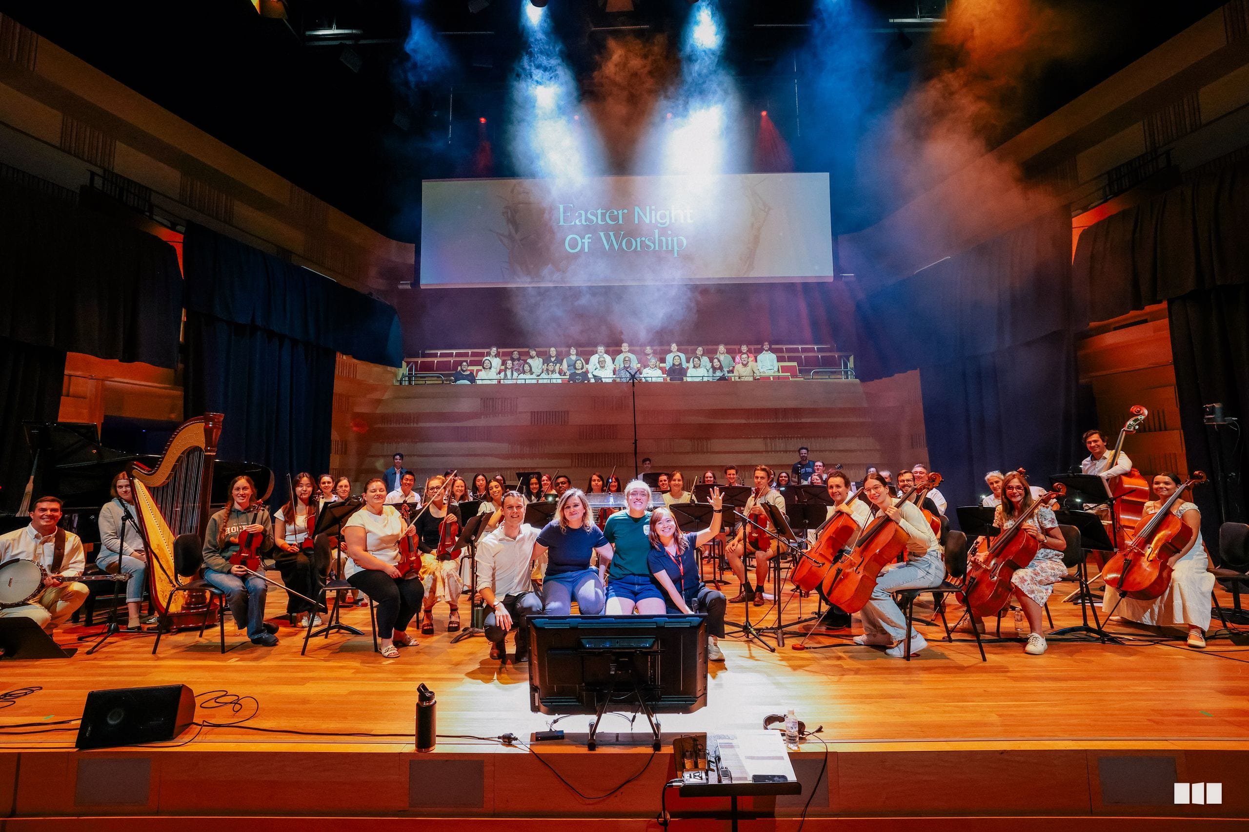 A large symphony orchestra and choir perform together on stage during an Easter Night of Worship event, with dramatic blue and orange lighting and projection screens above.