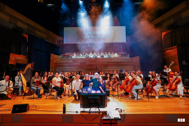 A large symphony orchestra and choir perform together on stage during an Easter Night of Worship event, with dramatic blue and orange lighting and projection screens above.