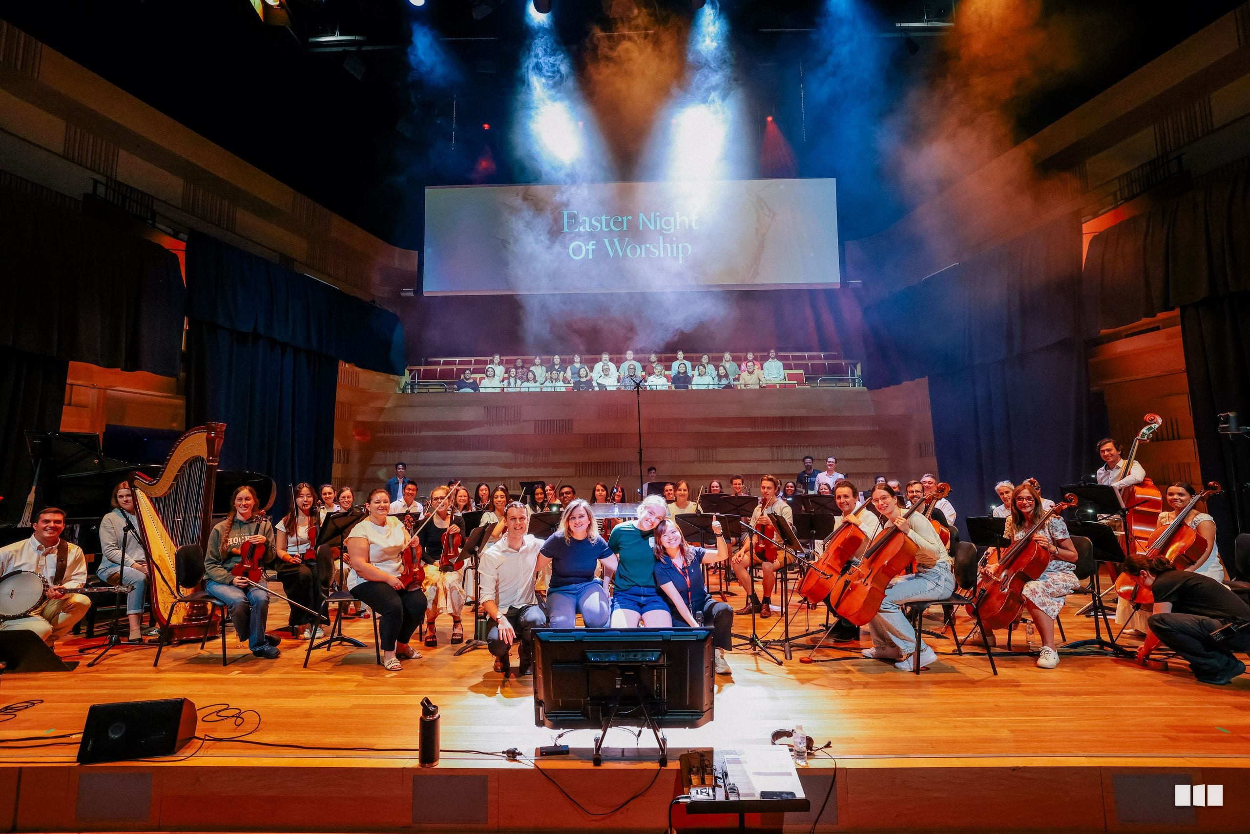 A full orchestra and choir perform together on stage during an Easter Night of Worship concert, with dramatic blue and orange lighting and a large projection screen above.