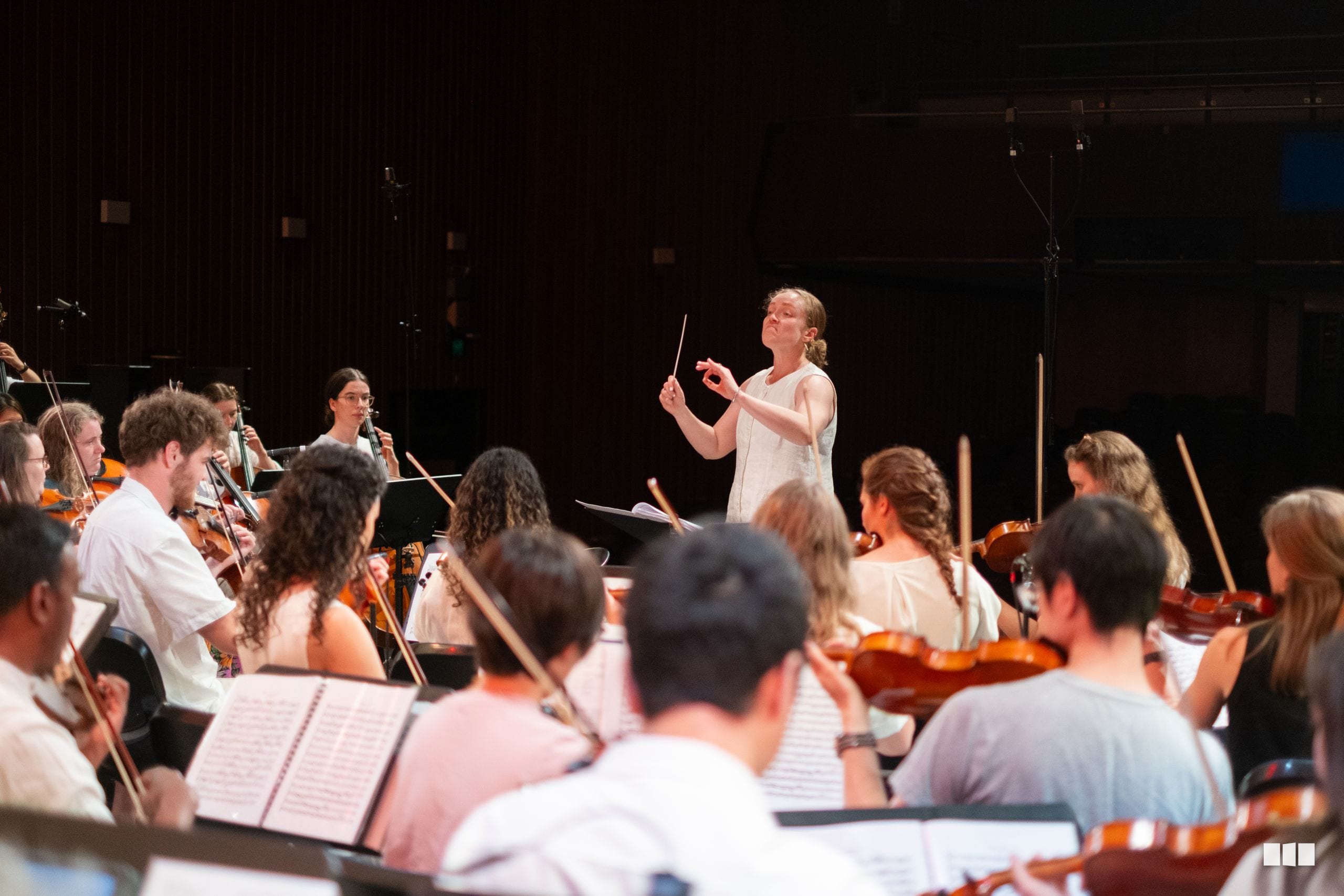 A conductor leads a string orchestra during rehearsal, with musicians holding violins and cellos while following sheet music.