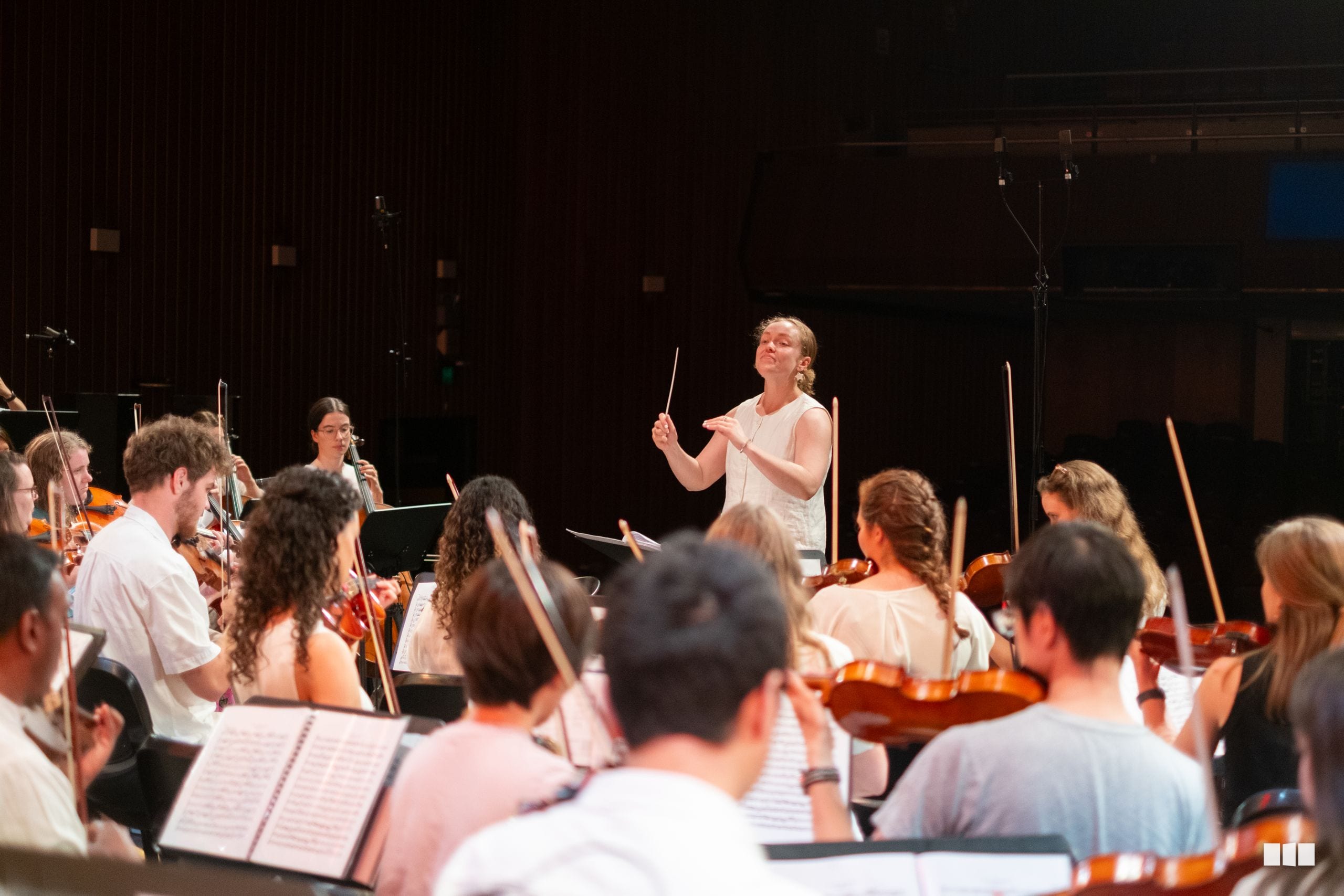 A conductor leads a string orchestra during rehearsal, with musicians holding violins and violas while reading from sheet music.