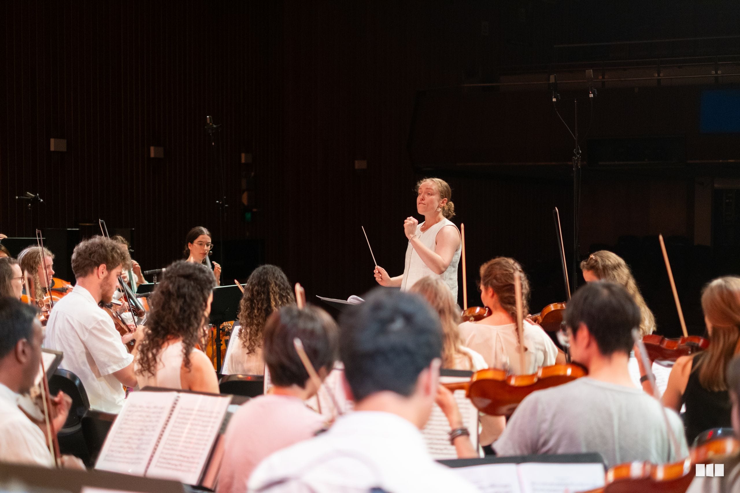 A conductor leads a string orchestra during a rehearsal or performance in a concert hall.