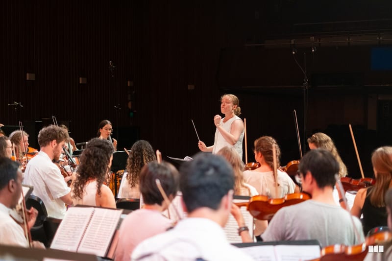 A conductor leads a string orchestra during a rehearsal or performance in a concert hall.