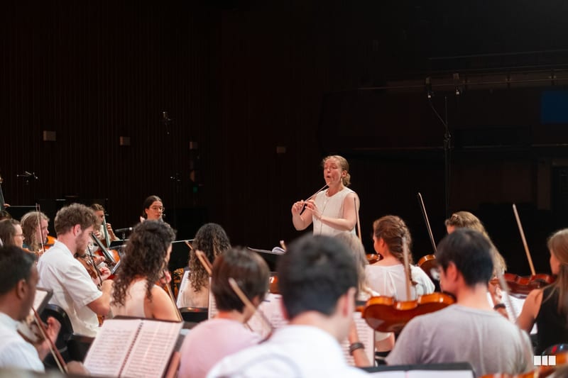 A conductor leads a string orchestra section during a rehearsal or performance on stage.