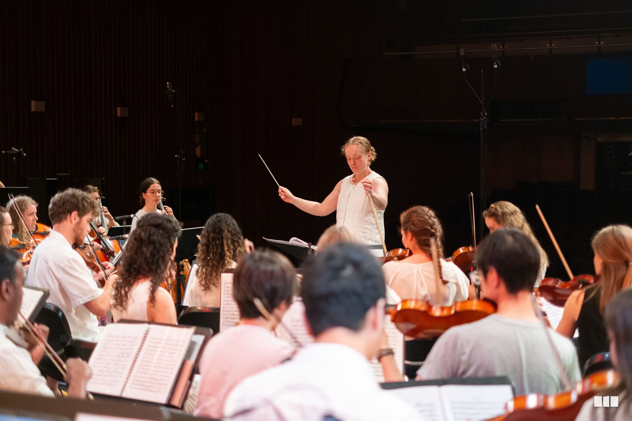 A conductor leads a string orchestra during rehearsal, with musicians holding violins and cellos while reading from sheet music.