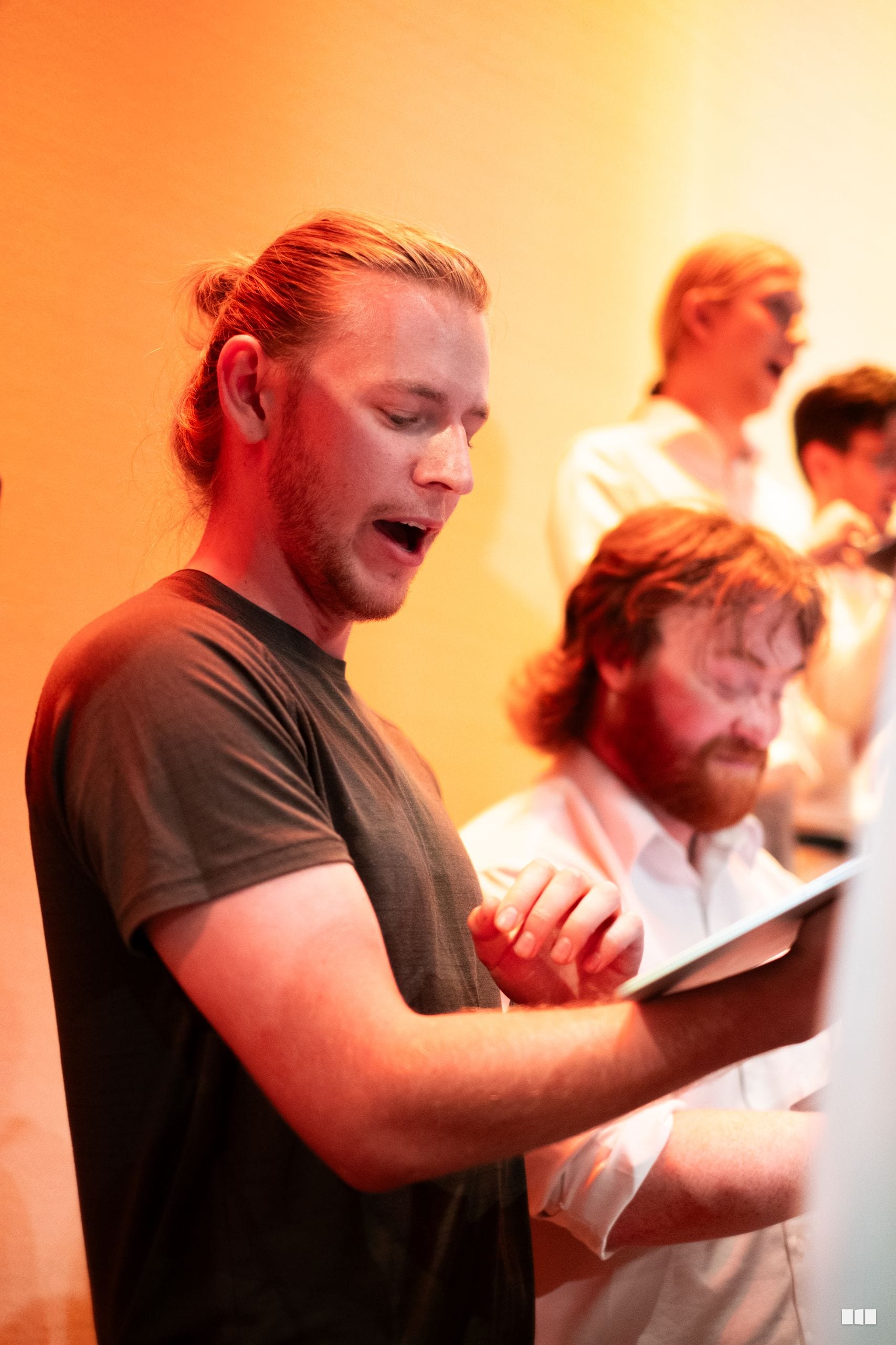 A male singer with red hair in a dark shirt performs with other choir members holding sheet music under warm stage lighting.