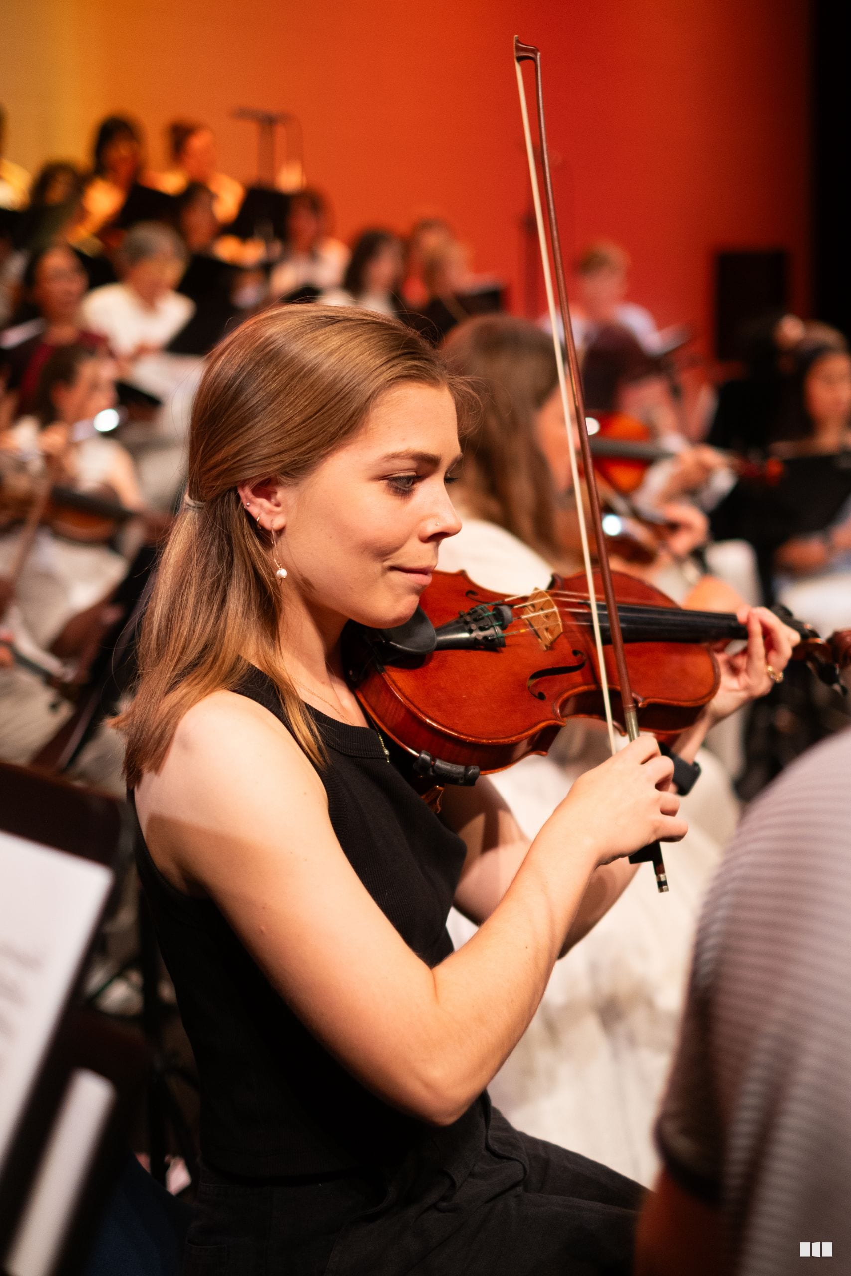 A young violinist performs in front of an engaged audience in an intimate concert setting with warm stage lighting.