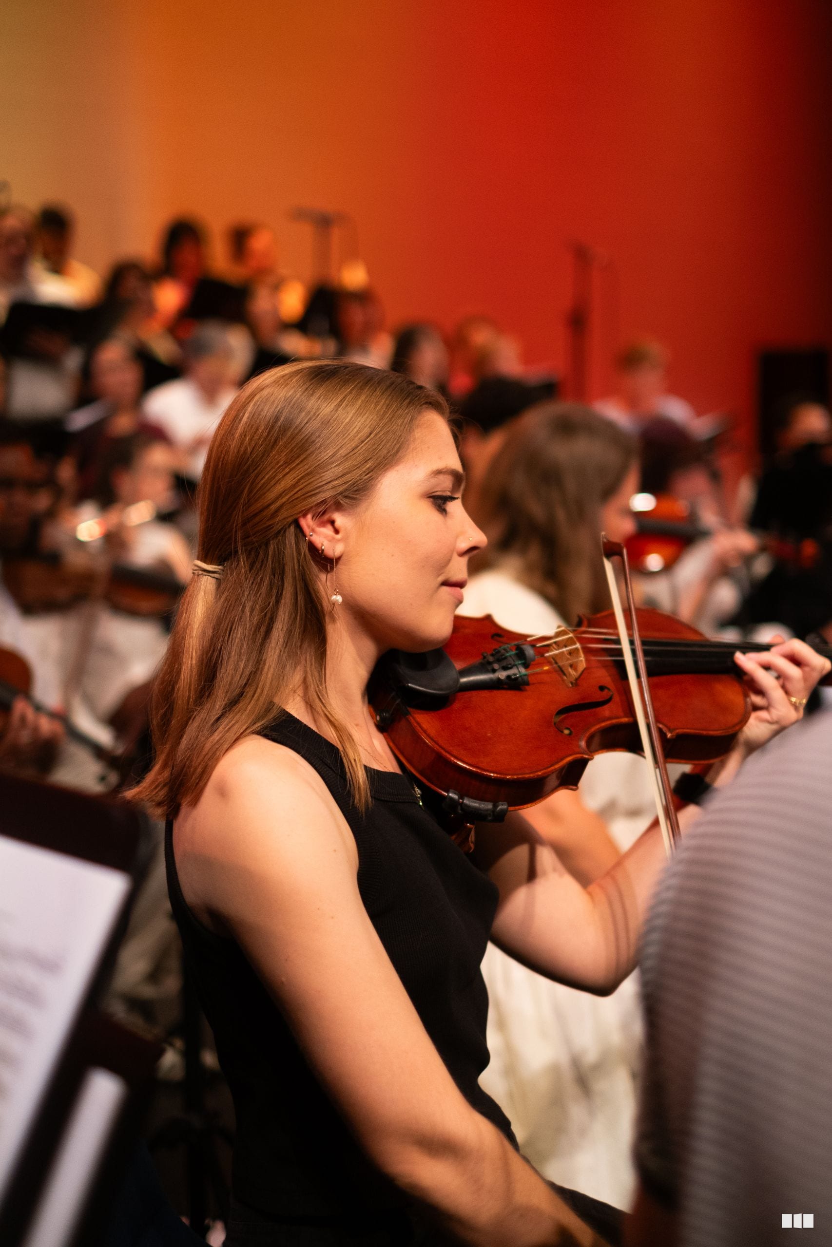 A young violinist in formal black attire performs with the orchestra during a concert against a warm-toned red backdrop.