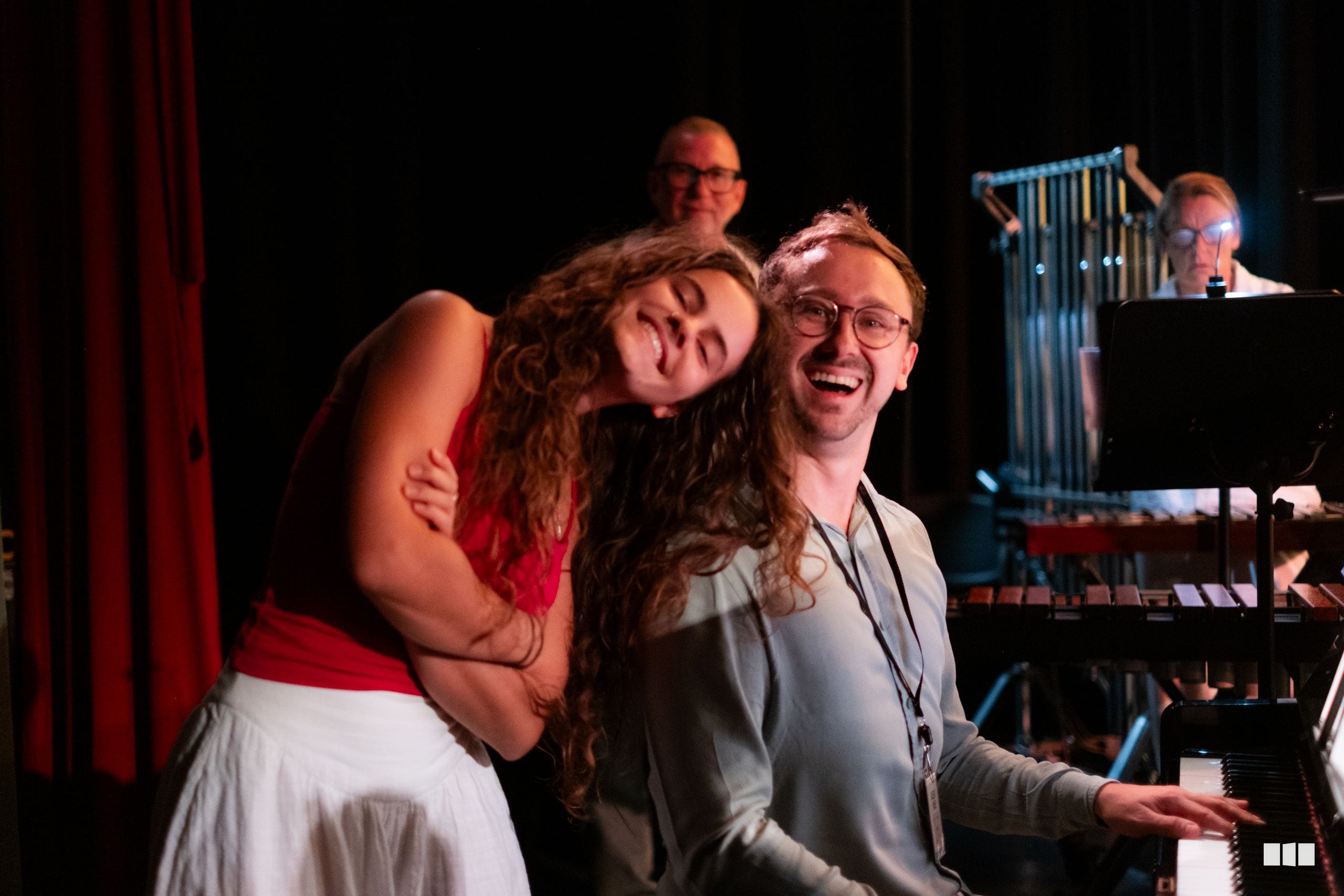 A pianist and soprano vocalist share an joyful moment during a performance, with orchestra members visible in the background.