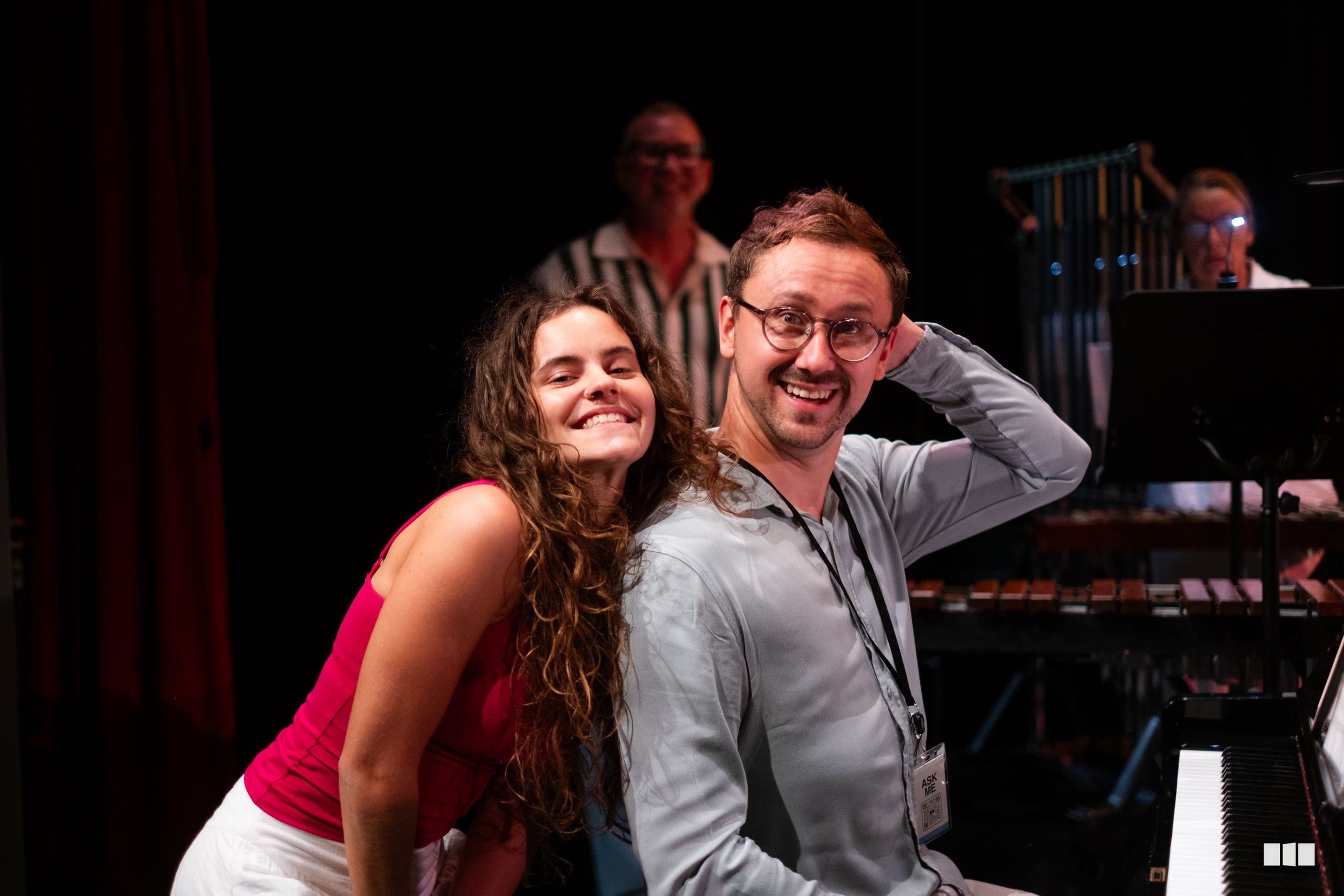 A young woman in a pink top and a man wearing glasses and a lanyard smile together on stage near a piano, with other musicians visible in the background.