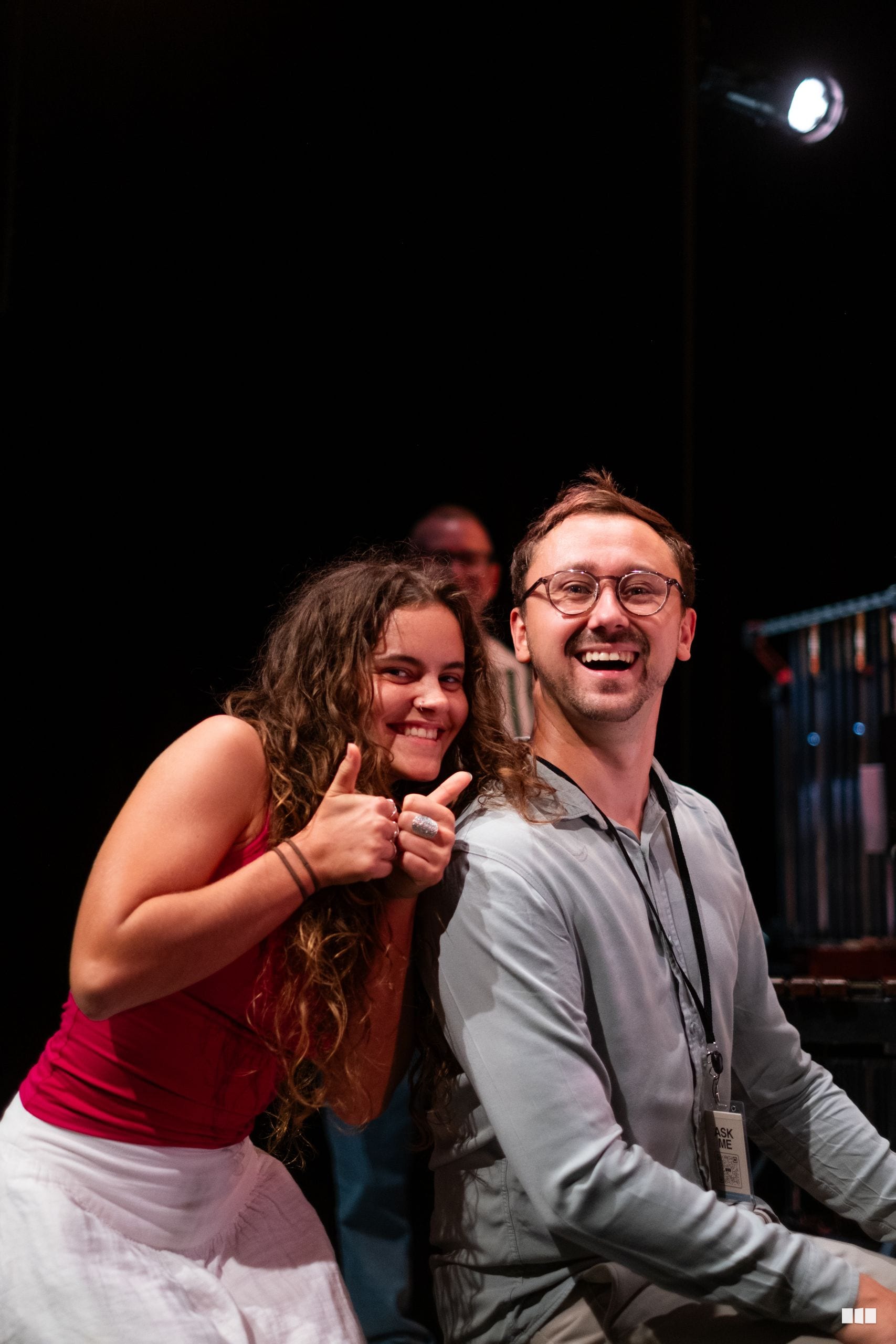 Two people smile and give thumbs up gestures at an evening event with stage lighting.