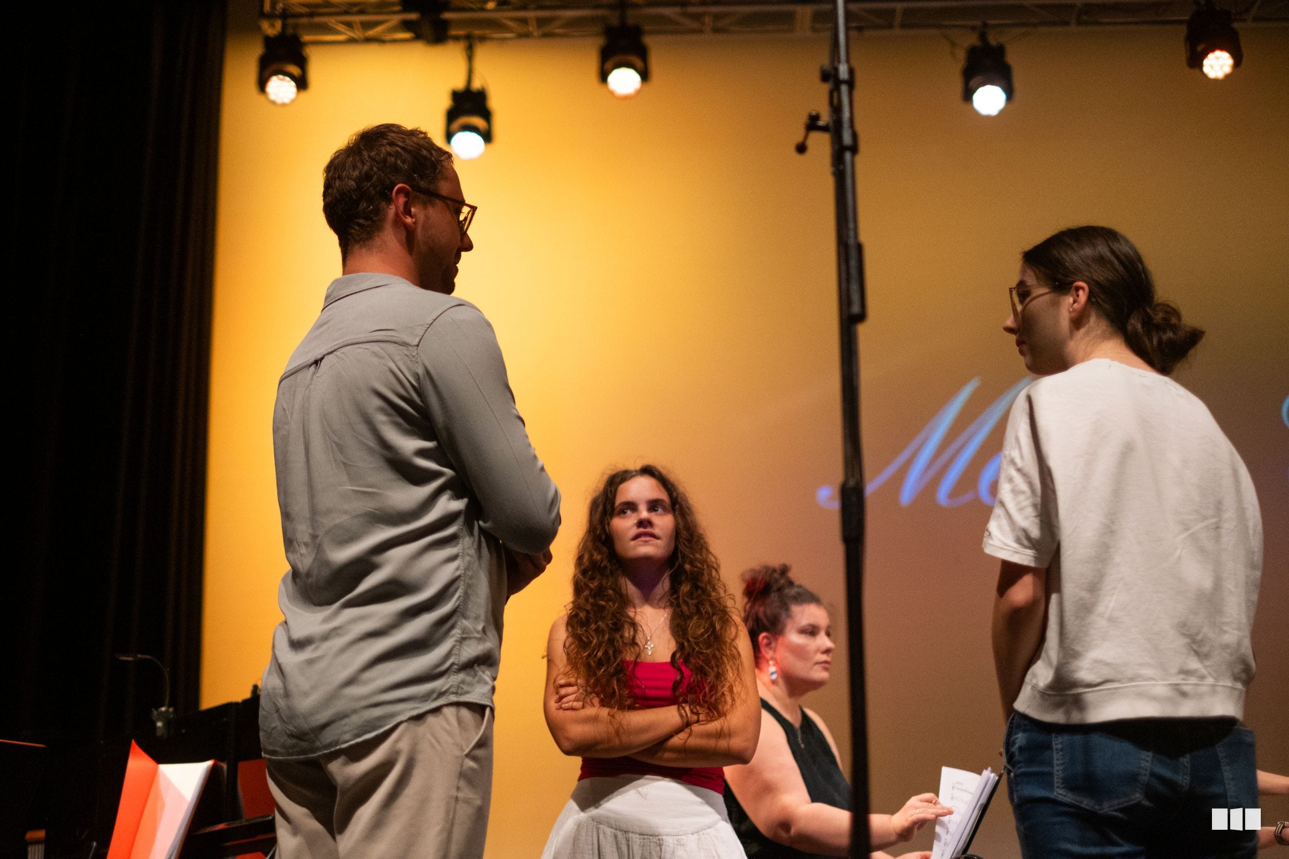 A group of young performers engage in discussion during a rehearsal or workshop on stage under warm lighting.