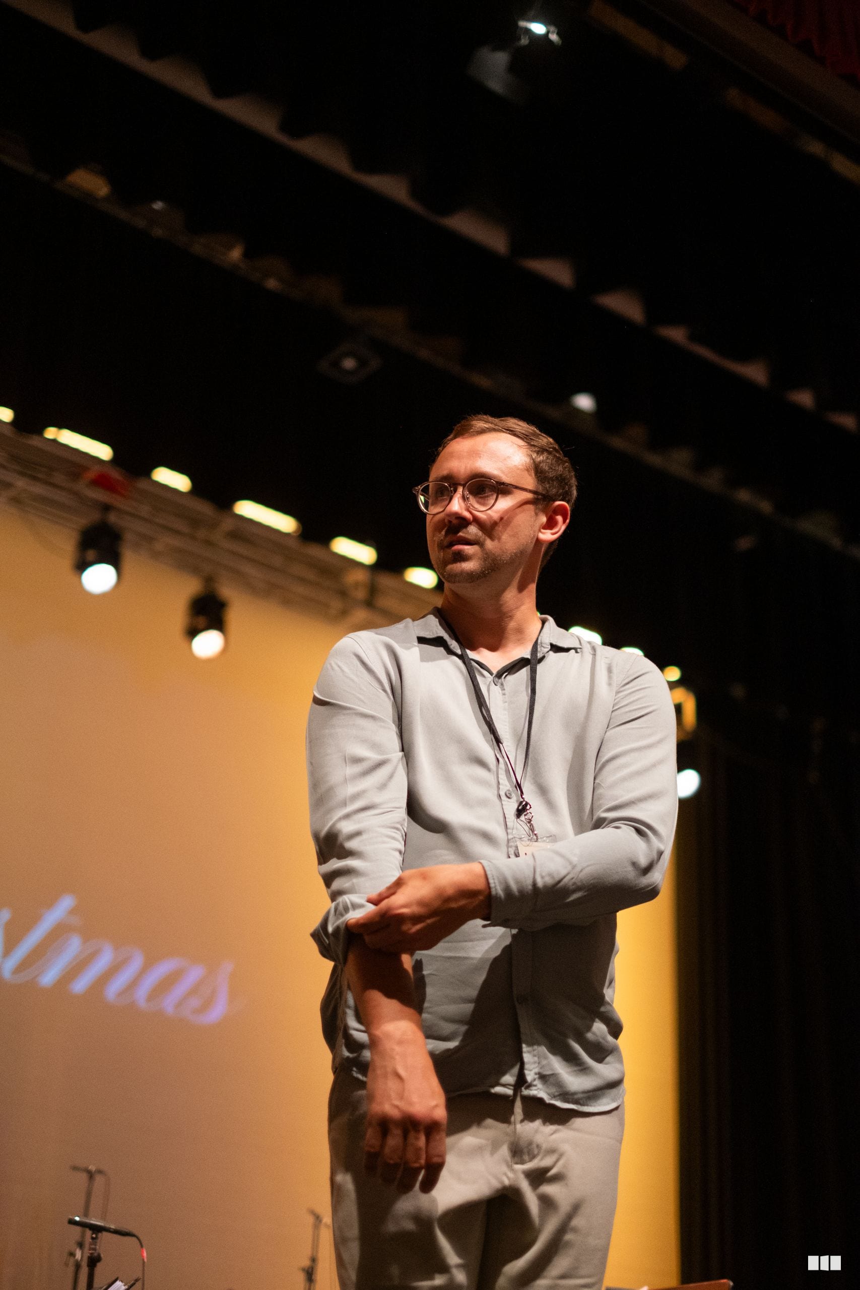 A man wearing glasses and a lanyard speaks on stage under warm lighting at an indoor venue.