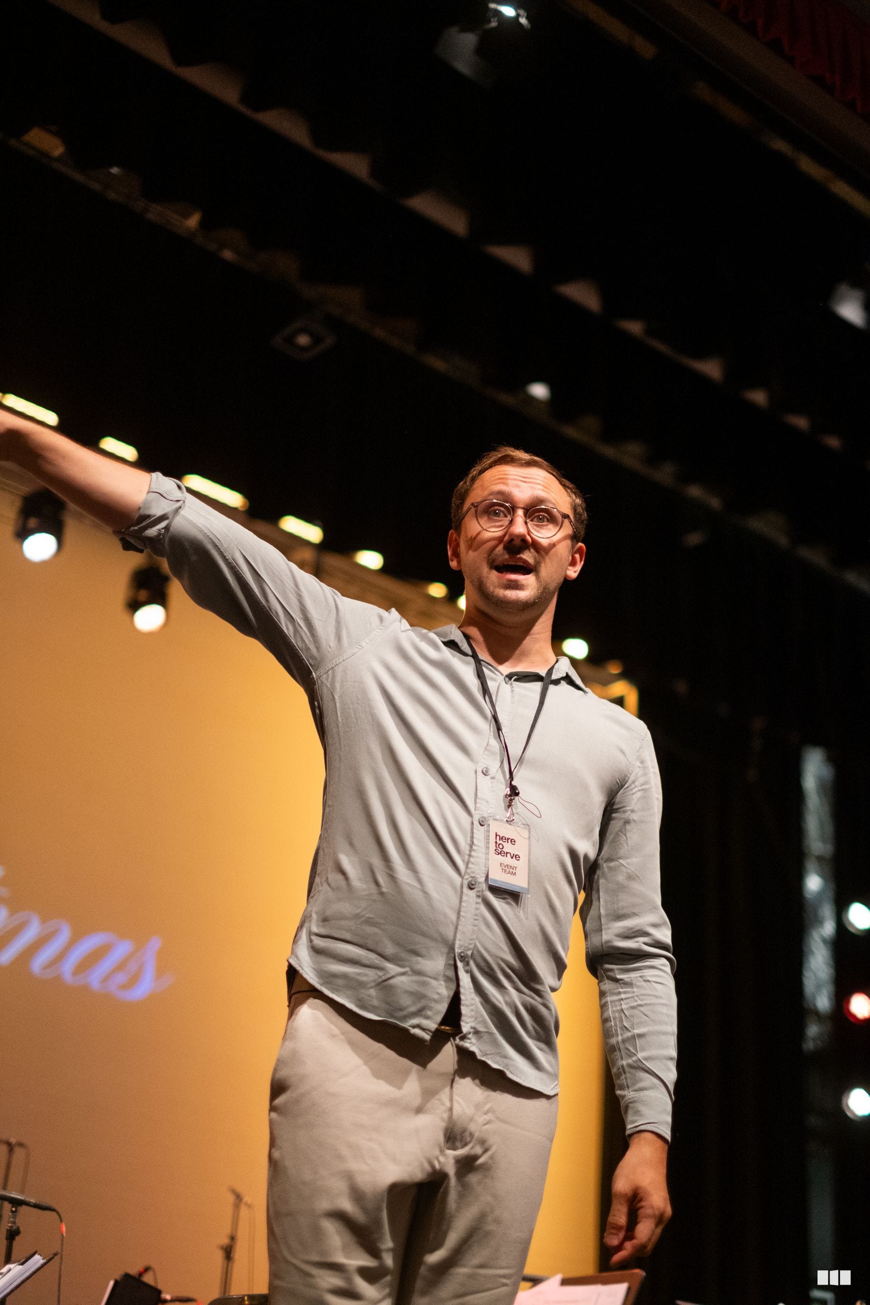 A speaker wearing glasses and a lanyard gestures while addressing an audience on stage with warm yellow lighting.