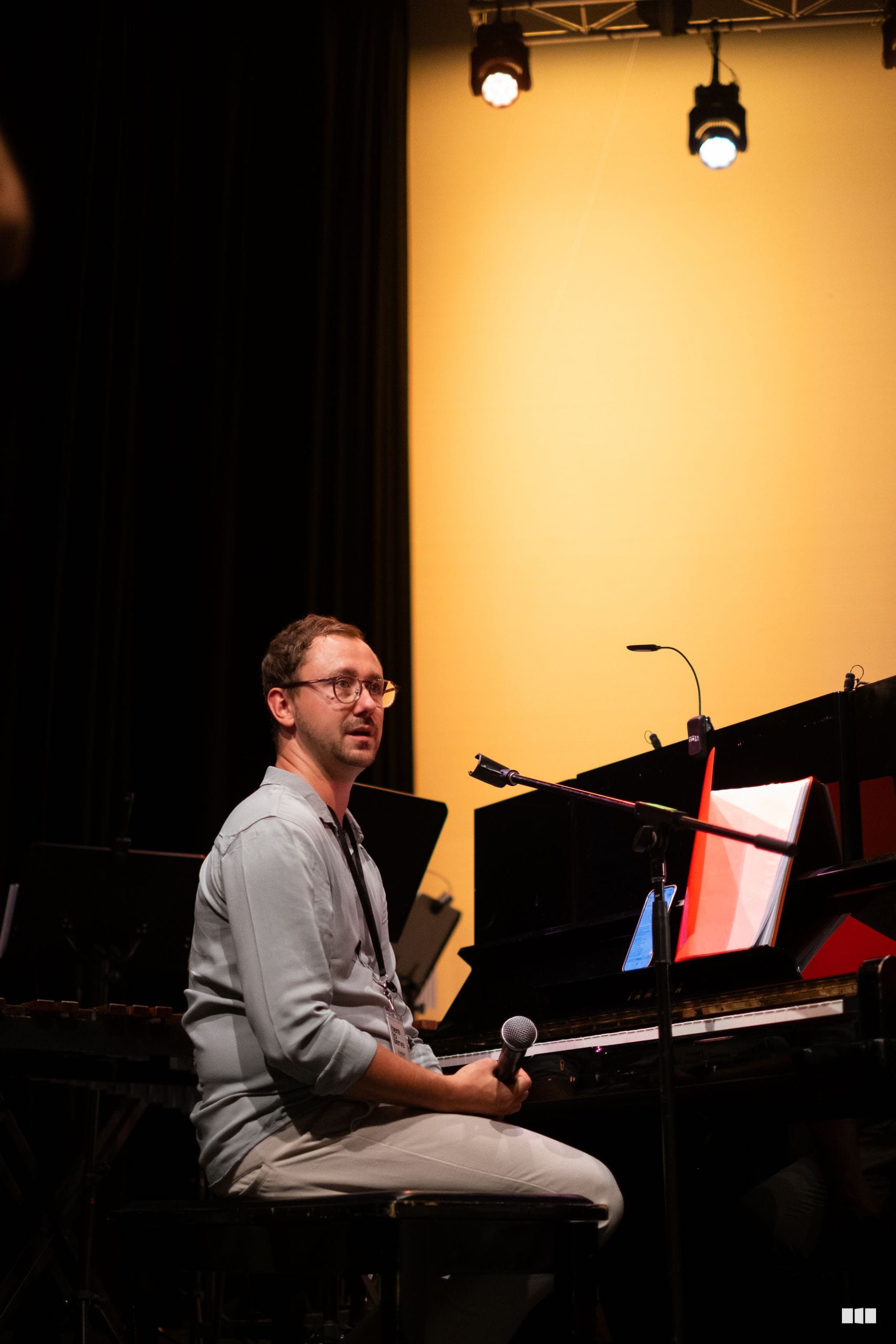 A male musician sits at a grand piano holding a microphone, speaking to the audience during a performance in a professionally lit theater setting.