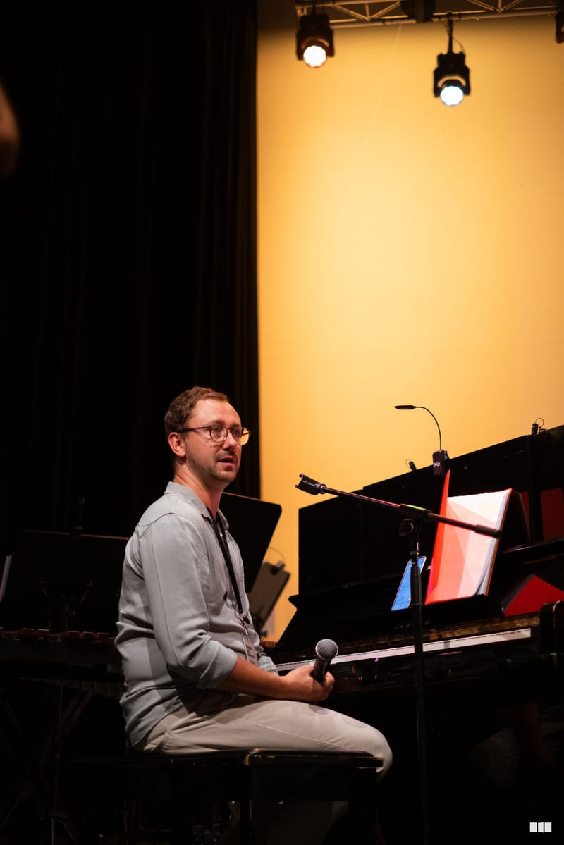 A male musician sits at a grand piano holding a microphone, speaking to the audience during a performance in a professionally lit theater setting.
