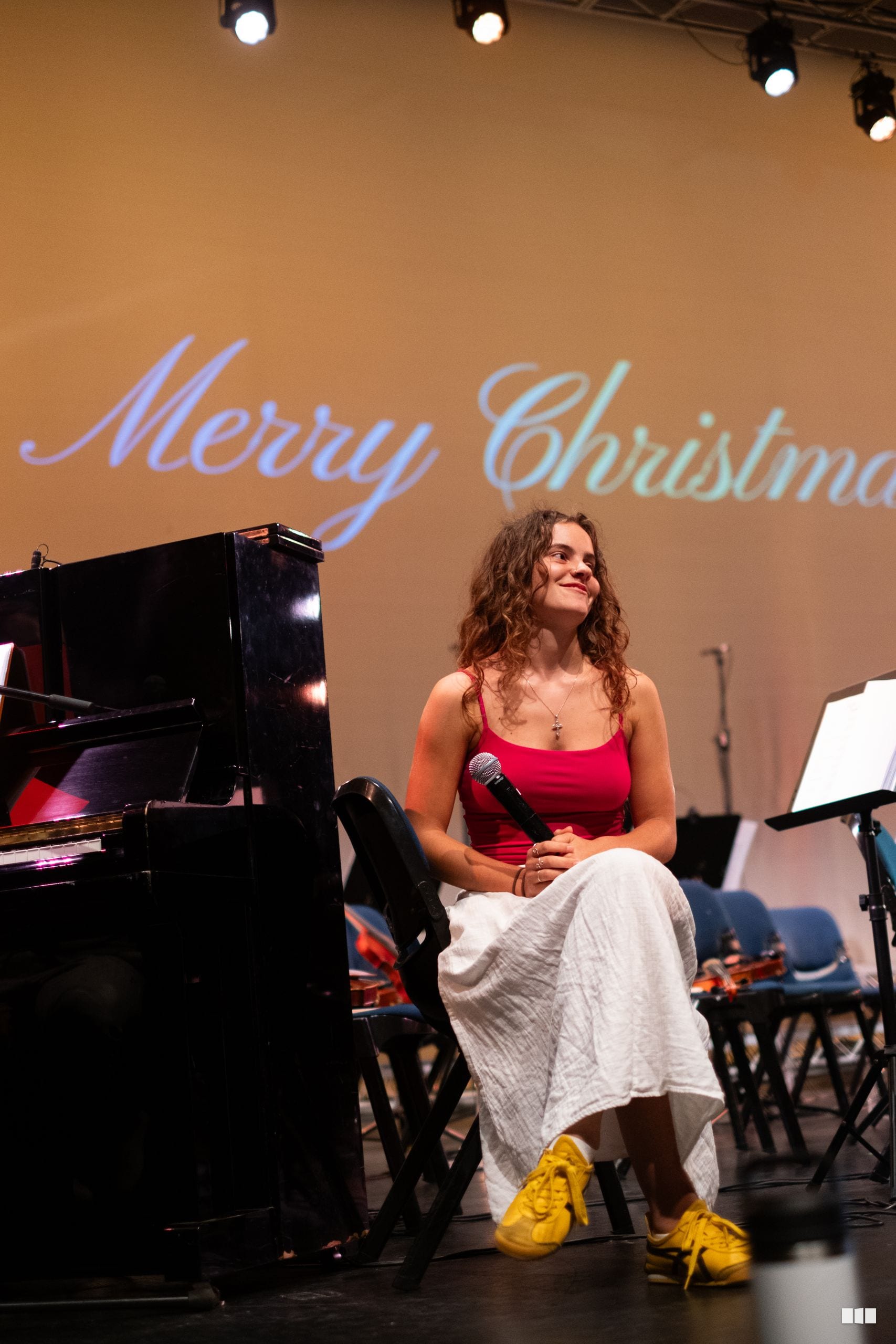 A female soloist in a red top and white skirt performs at the microphone beside a grand piano during a Christmas concert.