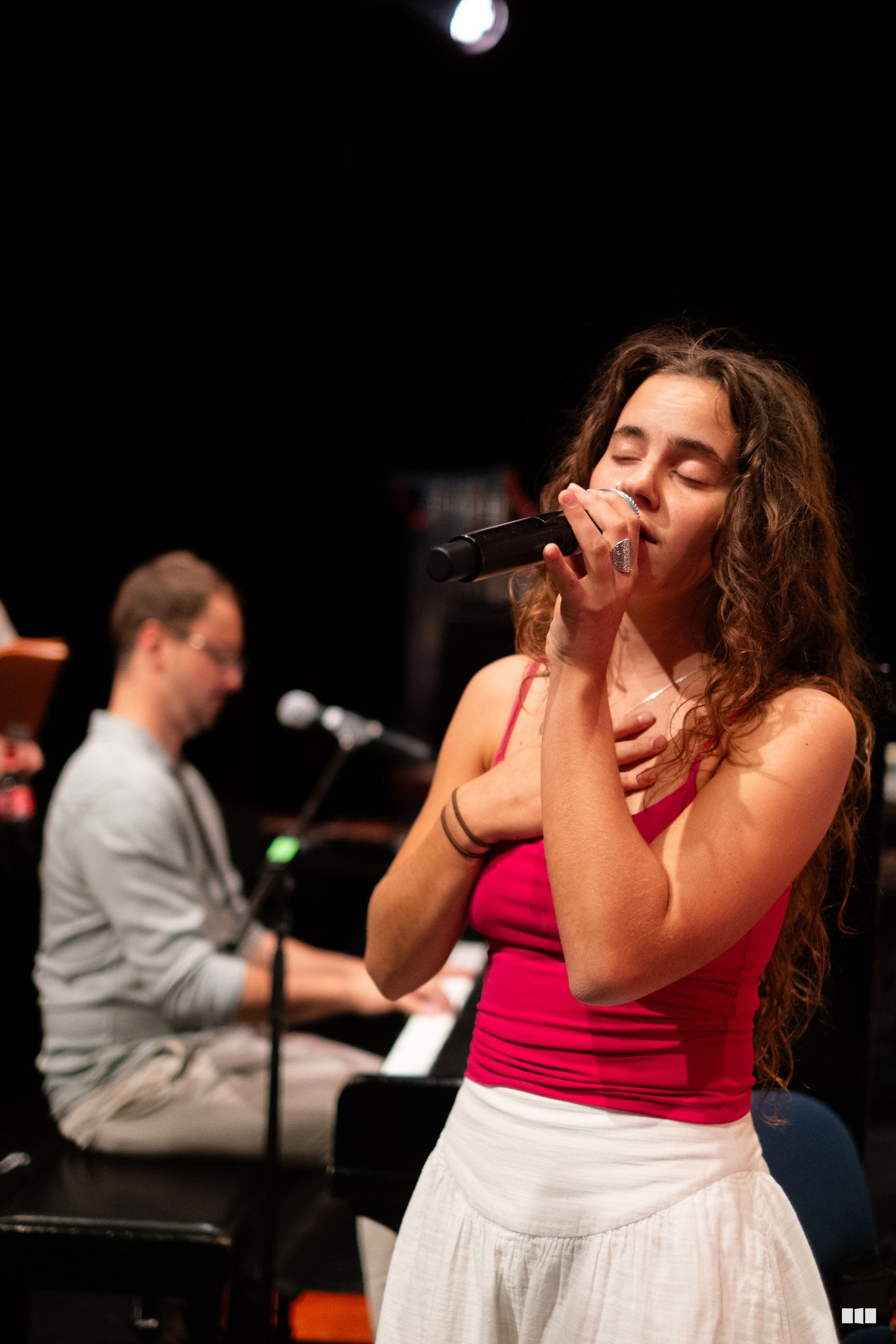 Female soloist singing into a microphone with a pianist accompanying her on stage during a performance.